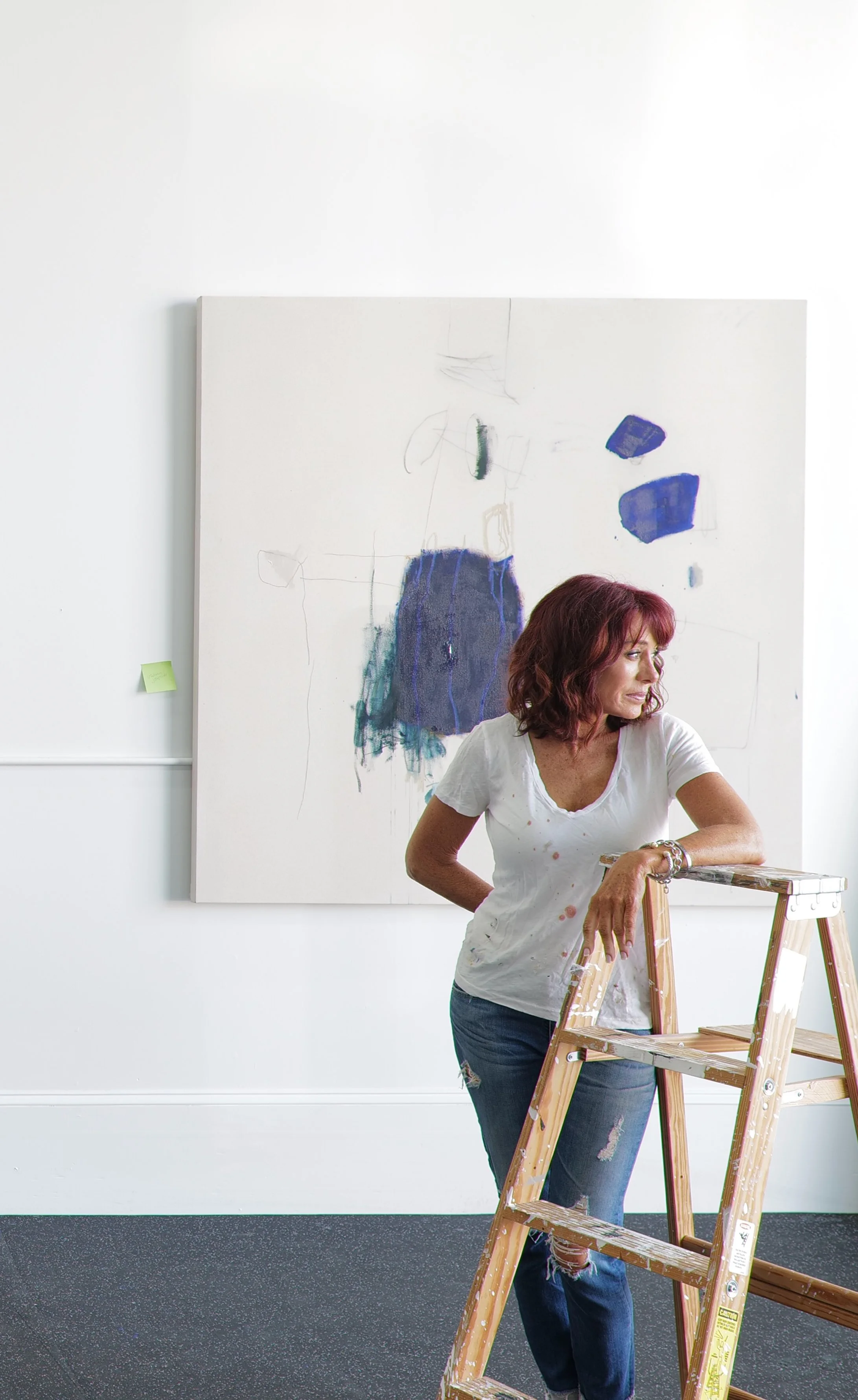 Chris Brandell, intuitive abstract artist with red hair standing next to a ladder in front of a painting.
