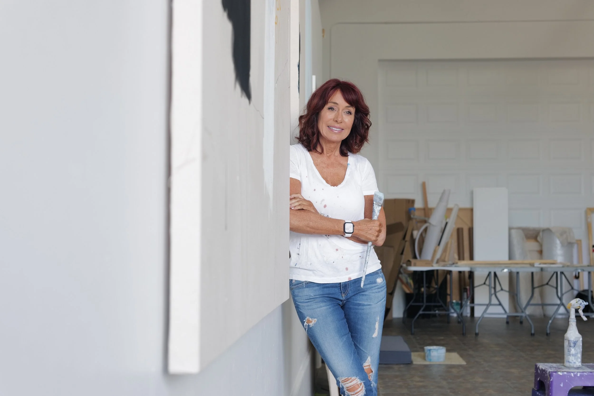 Chris Brandell, abstract artist, wearing a white splattered t-shirt and ripped jeans, standing in an art studio 7, holding a paintbrush, smiling, with art supplies and tables in the background.