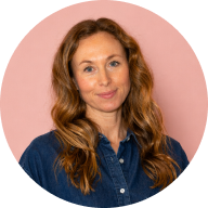 A woman with long, wavy red hair, wearing a blue denim shirt, smiling against a pink background.