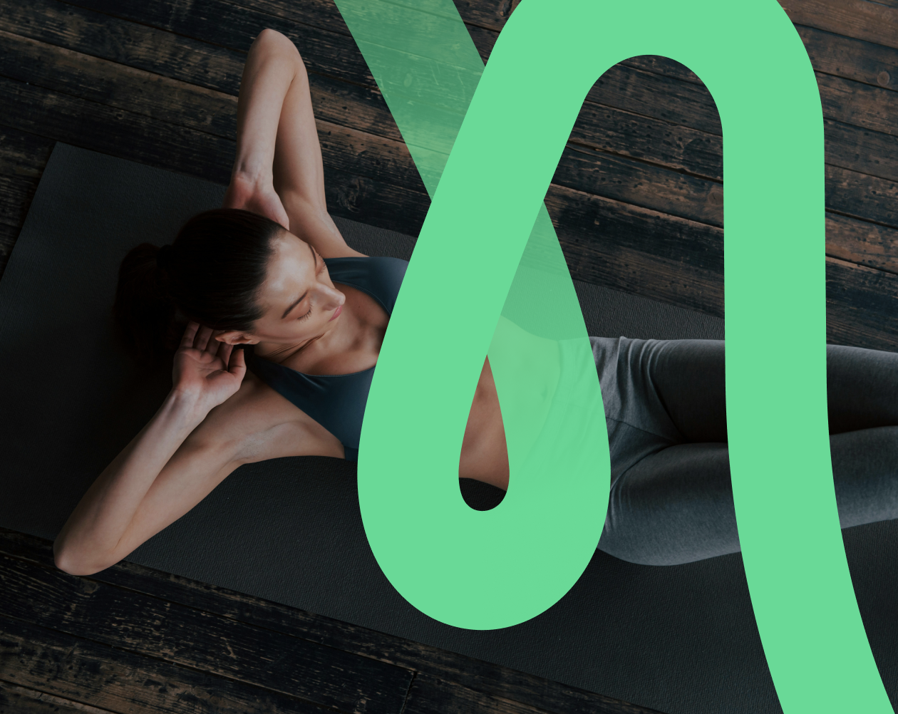 A woman lying on a yoga mat on a wooden floor, with her hands behind her head, doing a stretching exercise.