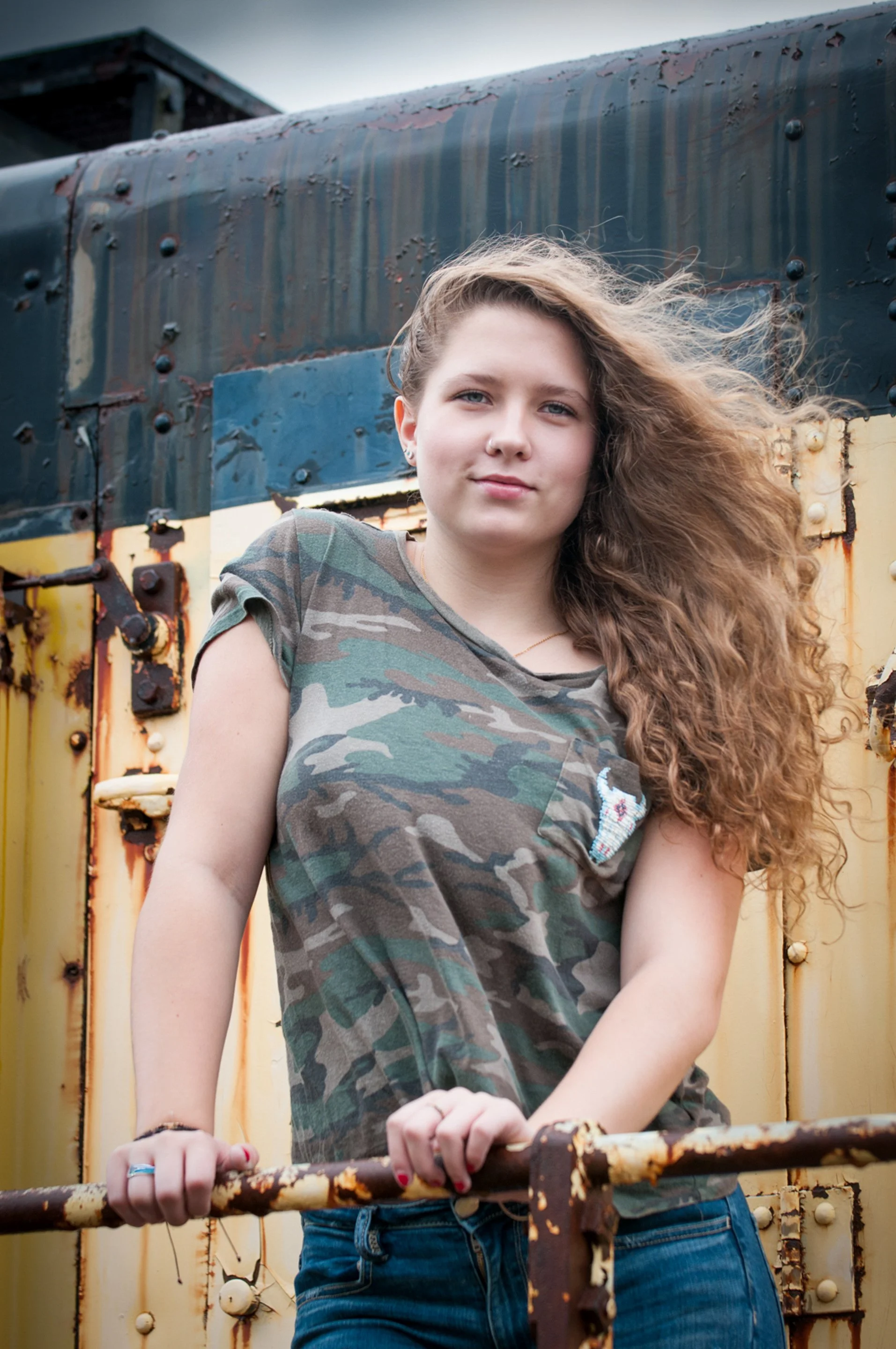 High school senior in camouflage tee leaning on a rusty yellow rail of an old train car, hair blowing in the wind.