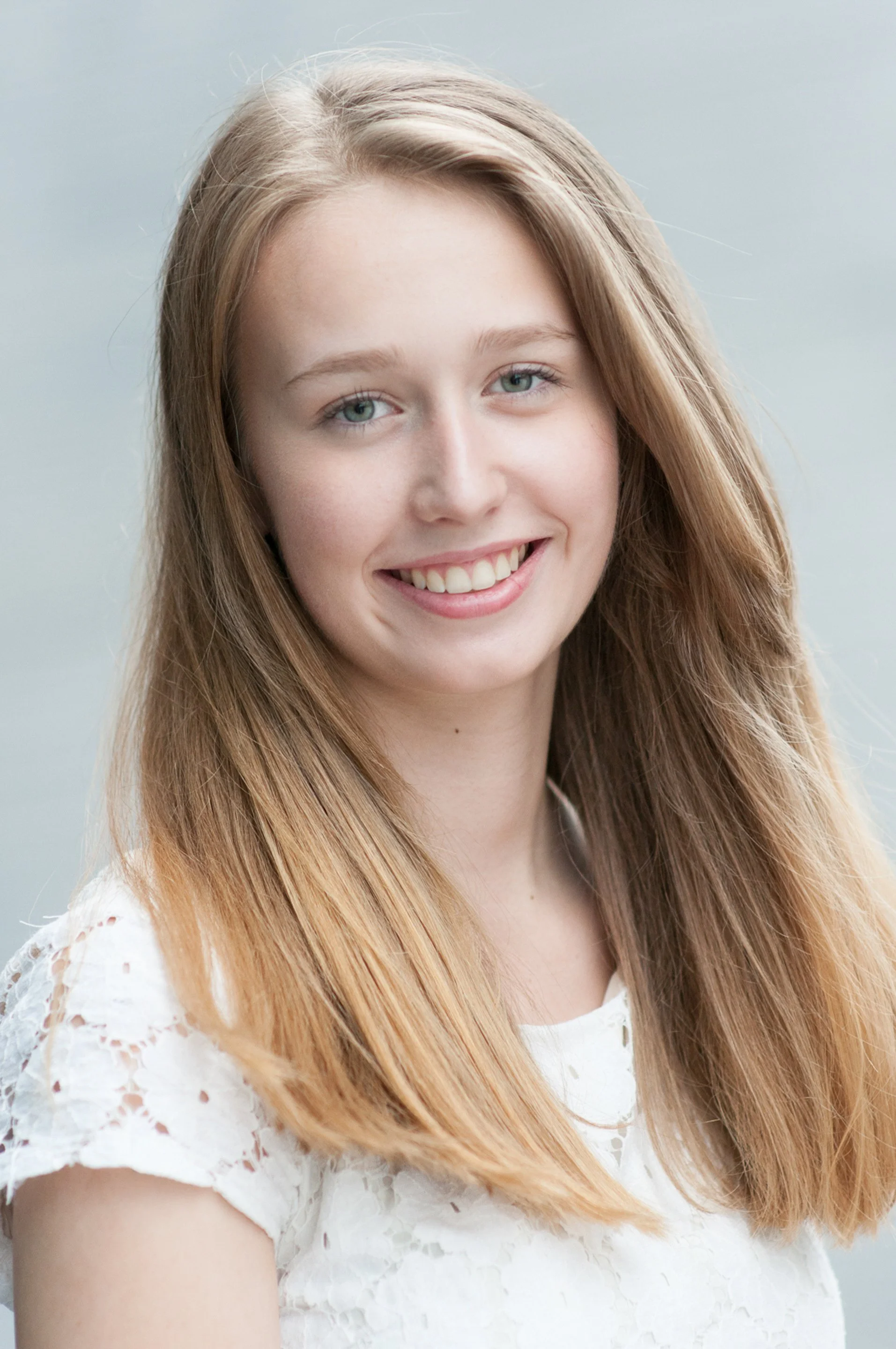 Senior portrait of a smiling girl in a white lace dress with along hair, gray backdrop