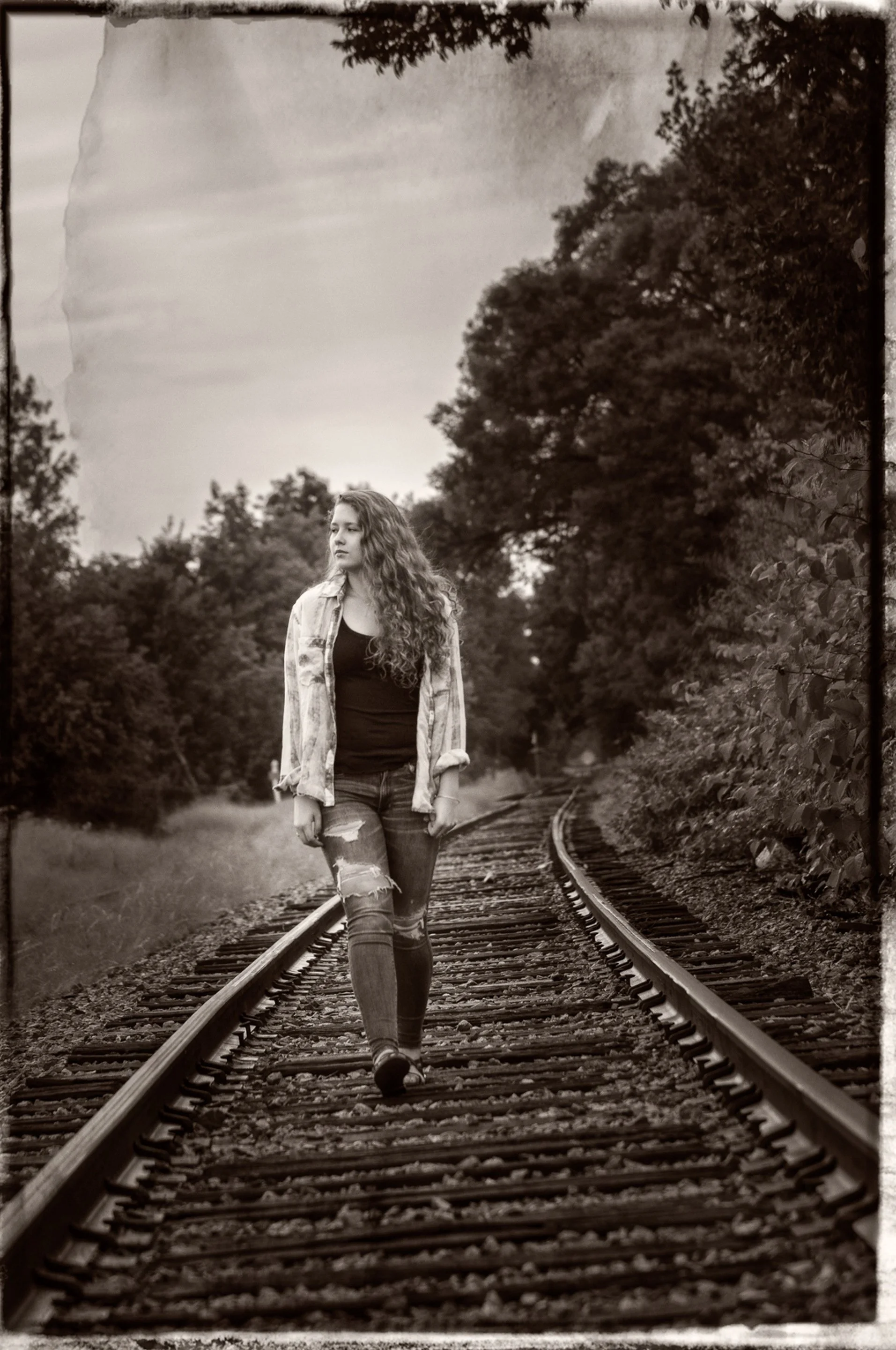 Sepia senior portrait o a girl in denim and a loose shirt walking down a curved railroad track
