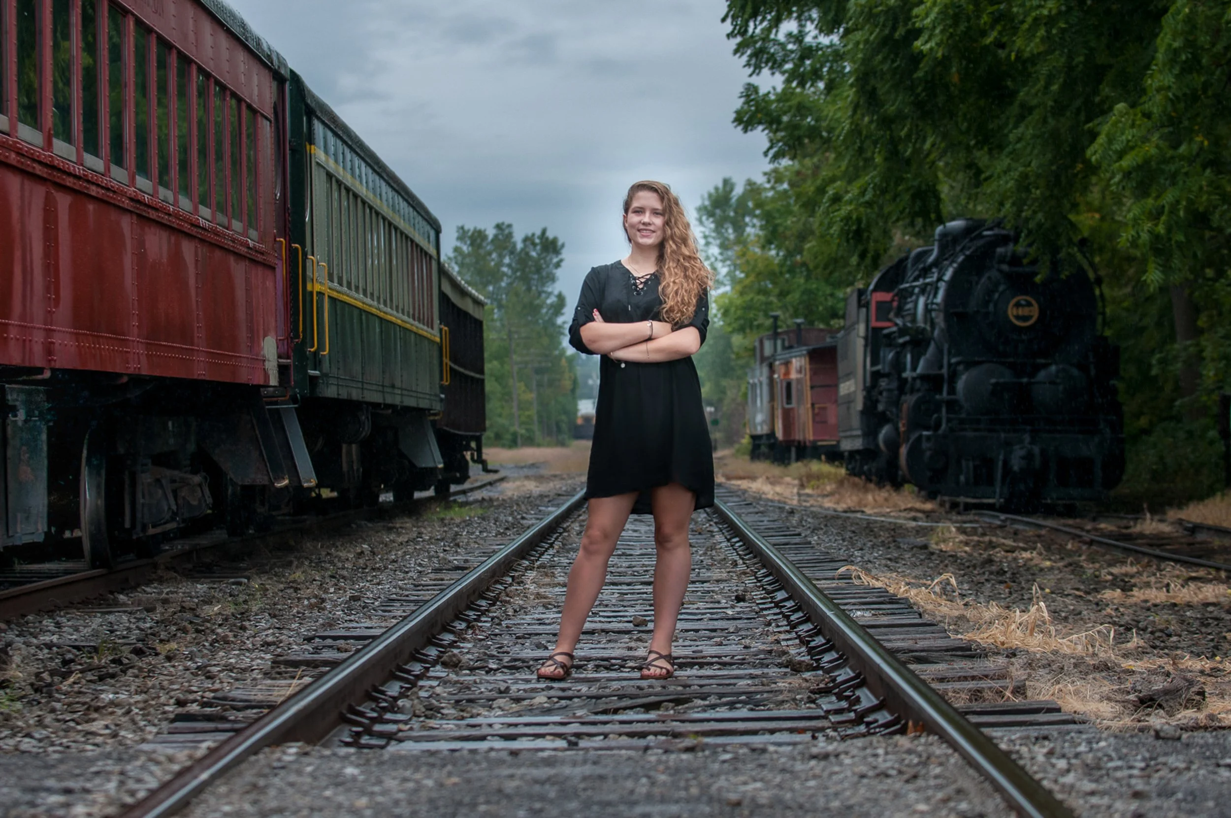 Senior in a black dress standing on railroad tracks with her arms crossed, vintage train cars on both sides.