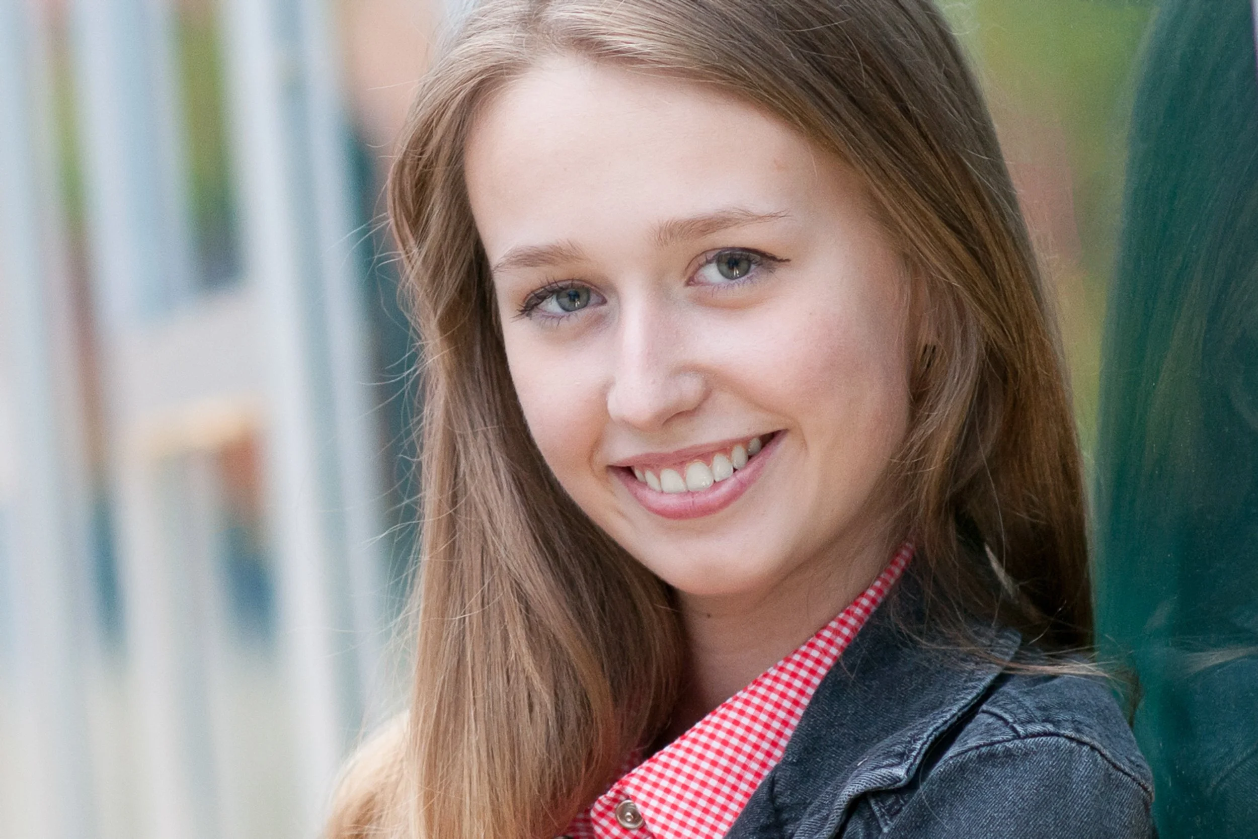 Close-up senior portrait of a smiling girl in a denim jacket and red gingham shirt.