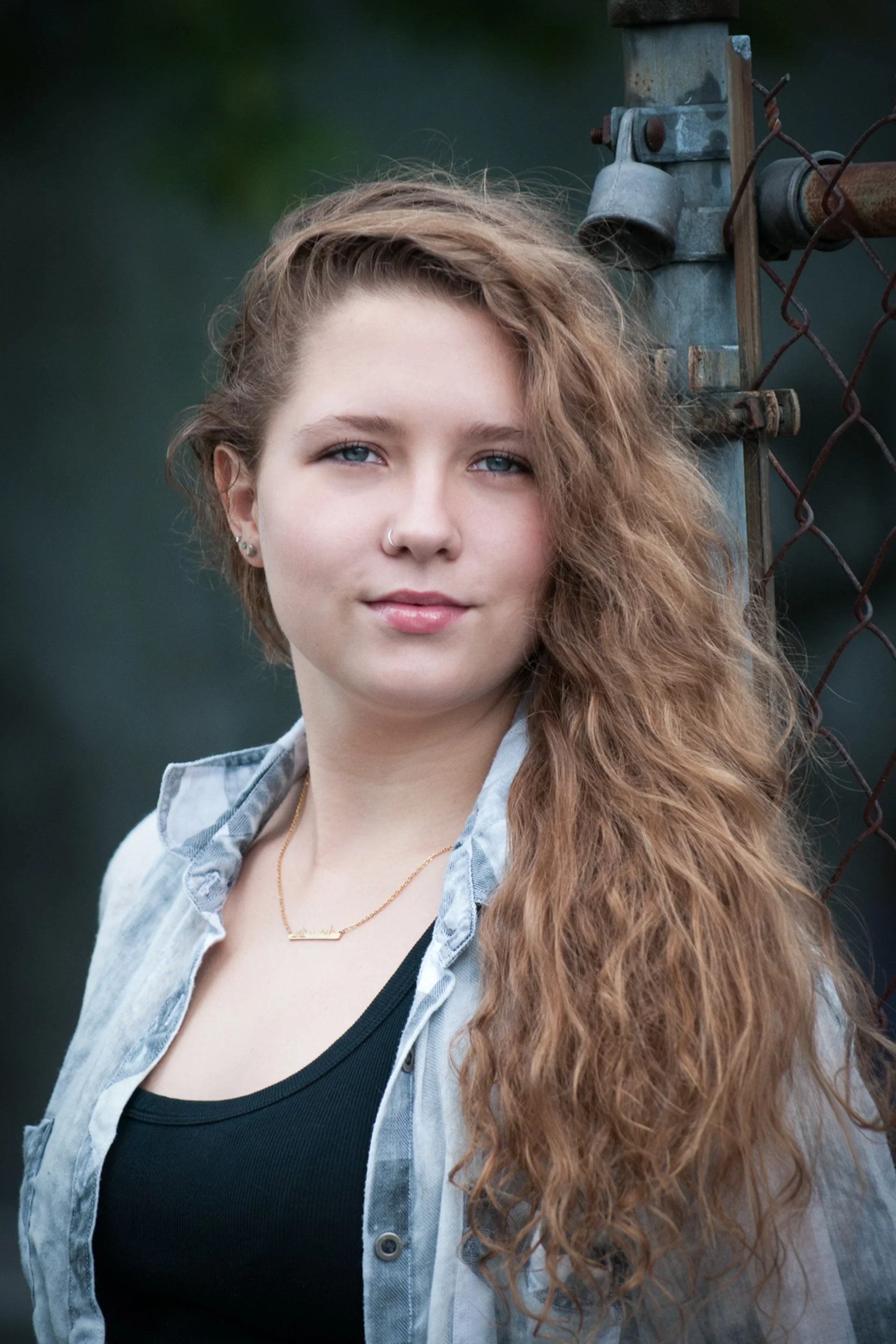 Close-up senior portrait o a girl in a denim shirt and black tank leaning against a chain-link fence.