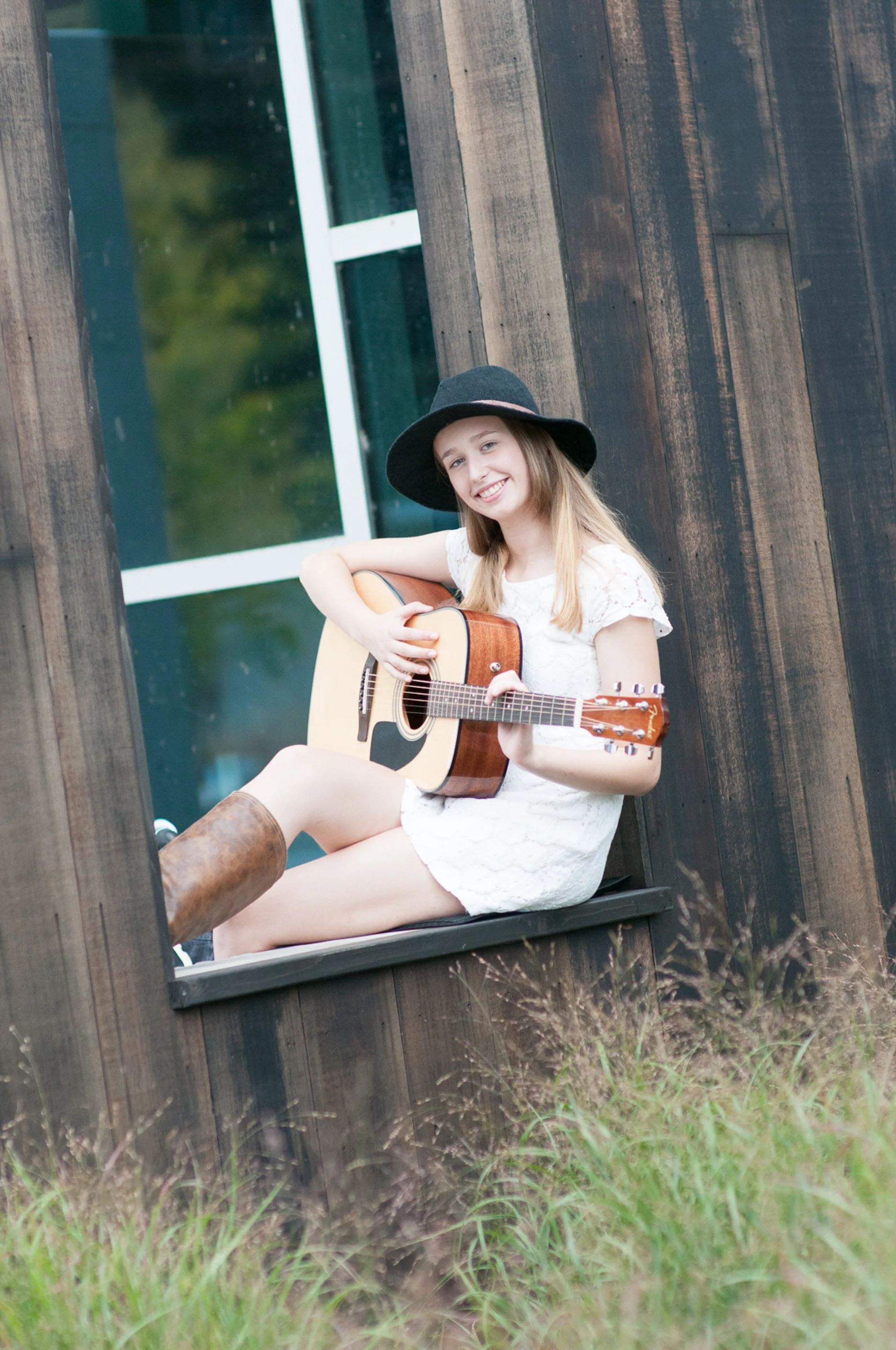 Senior sitting in a window seat playing an acoustic guitar, wearing a white lace dress, black hat, and brown boots.