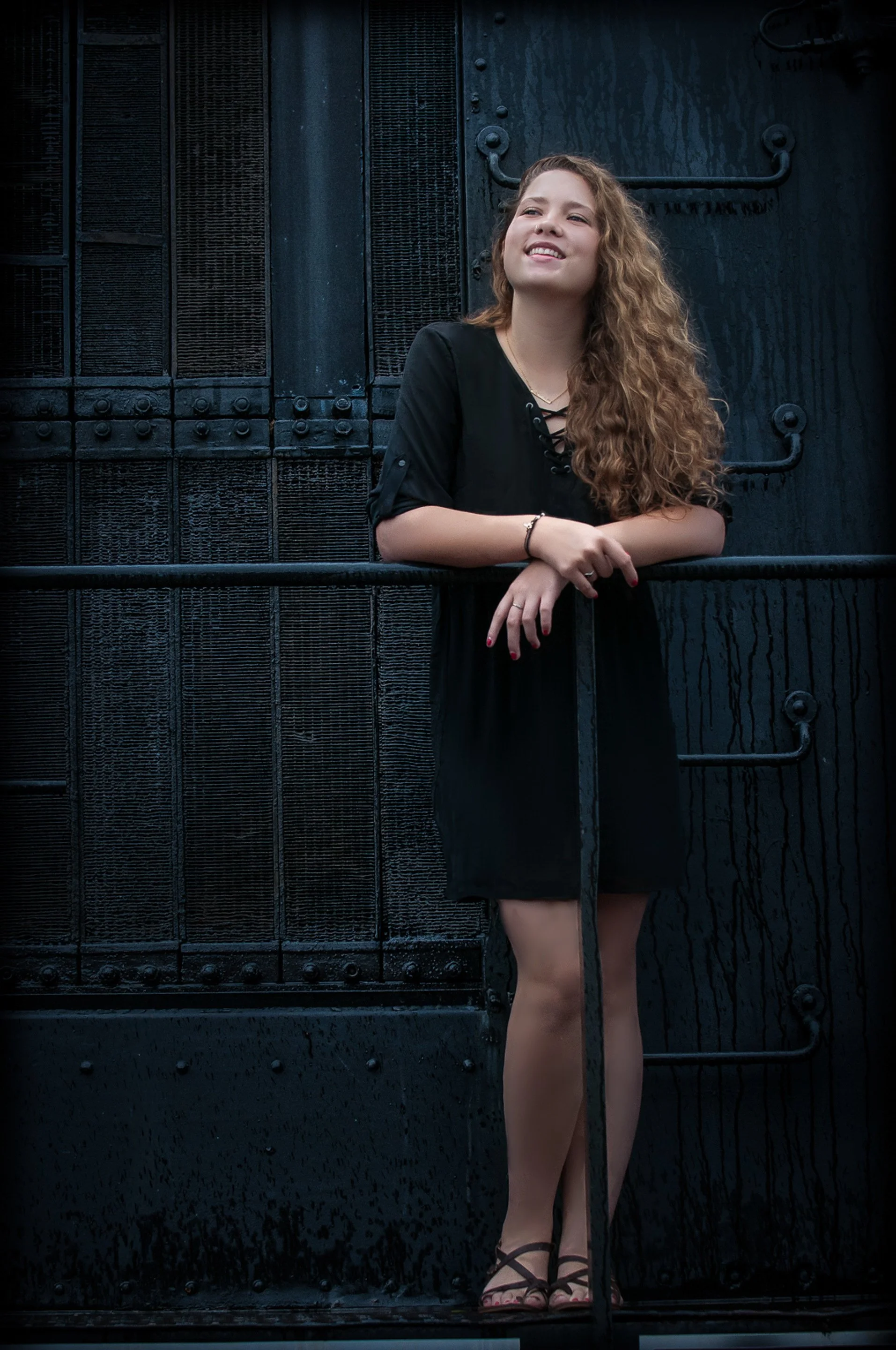 Senior in a black dress laughing and looking up as she leans on a rail in front of a dark train car.