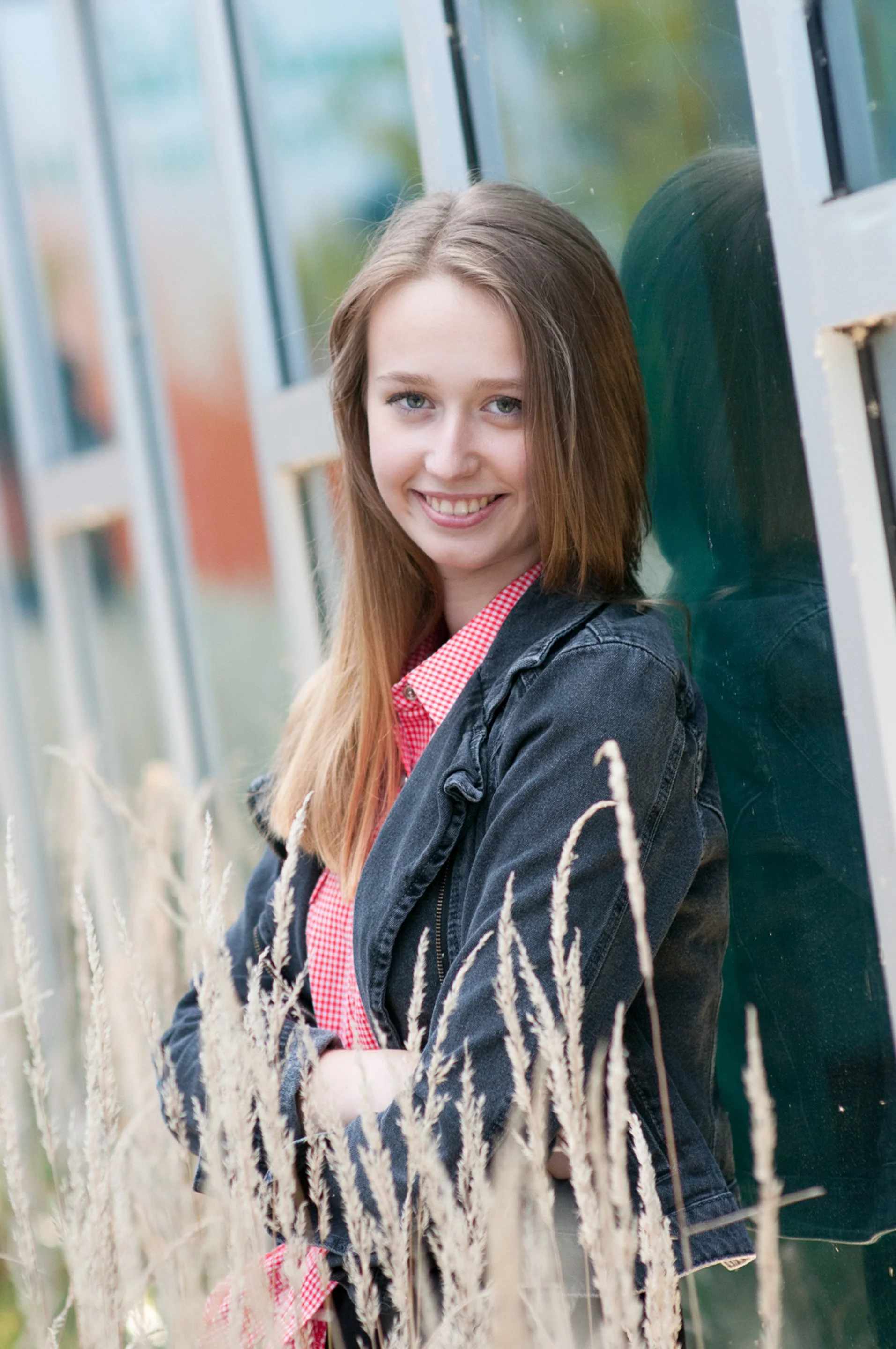 Senior leaning against a glass wall in a denim jacket and gingham shirt, tall grass in the foreground.
