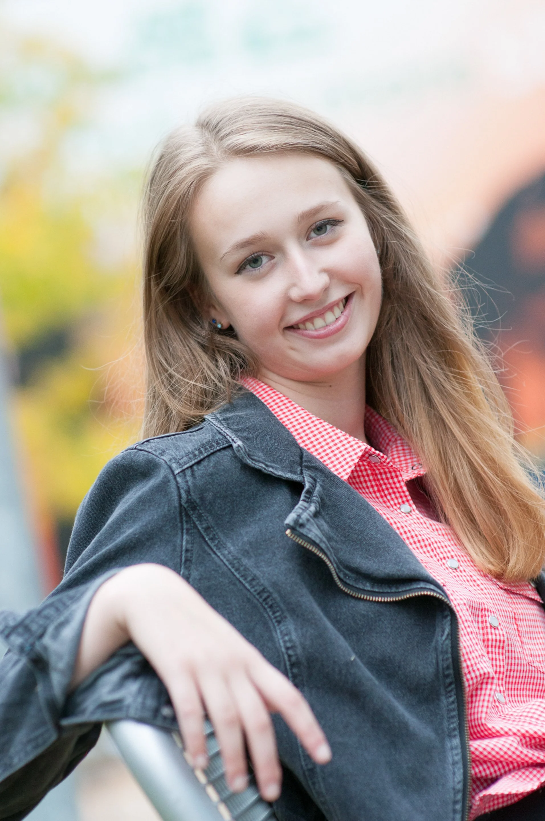 Senior smiling in a denim jacket and reg gingham shirt against a colorful blurred background
