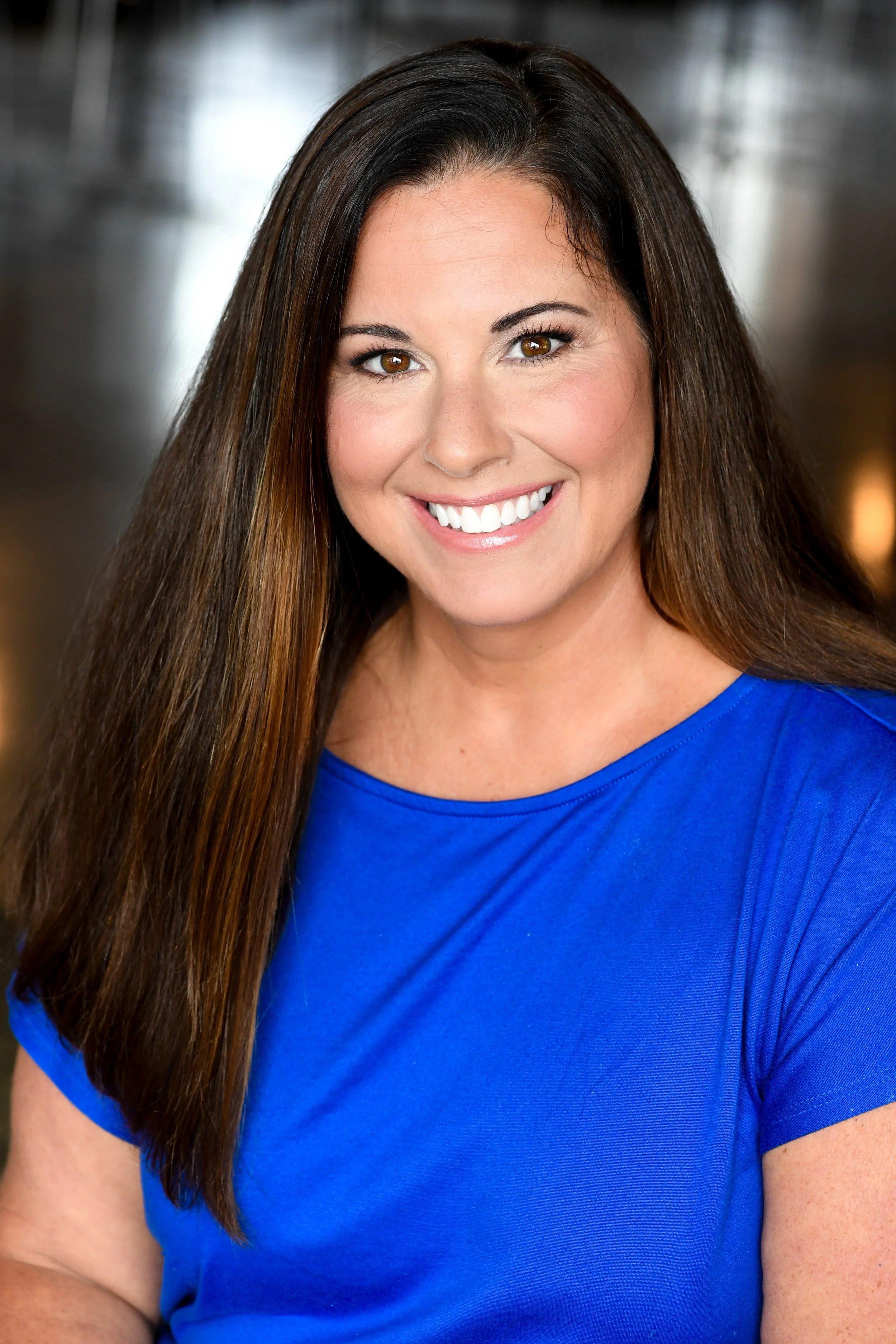 A woman with long brown hair smiling, wearing a bright blue top, in an indoor setting.