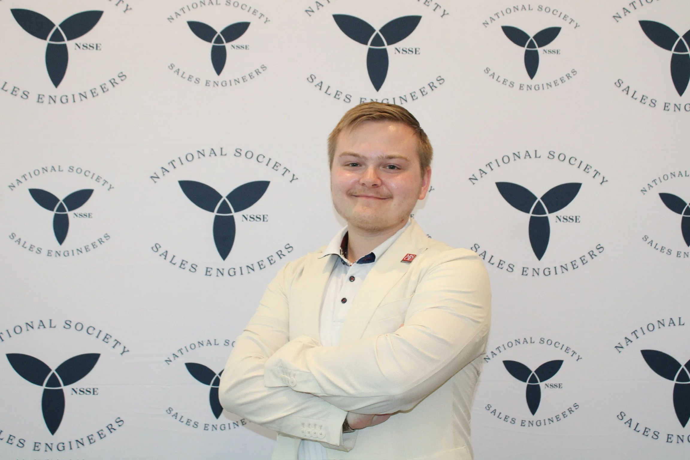 Young man with short light brown hair, blue eyes, wearing a gray plaid blazer and white shirt, smiling against a plain white background.