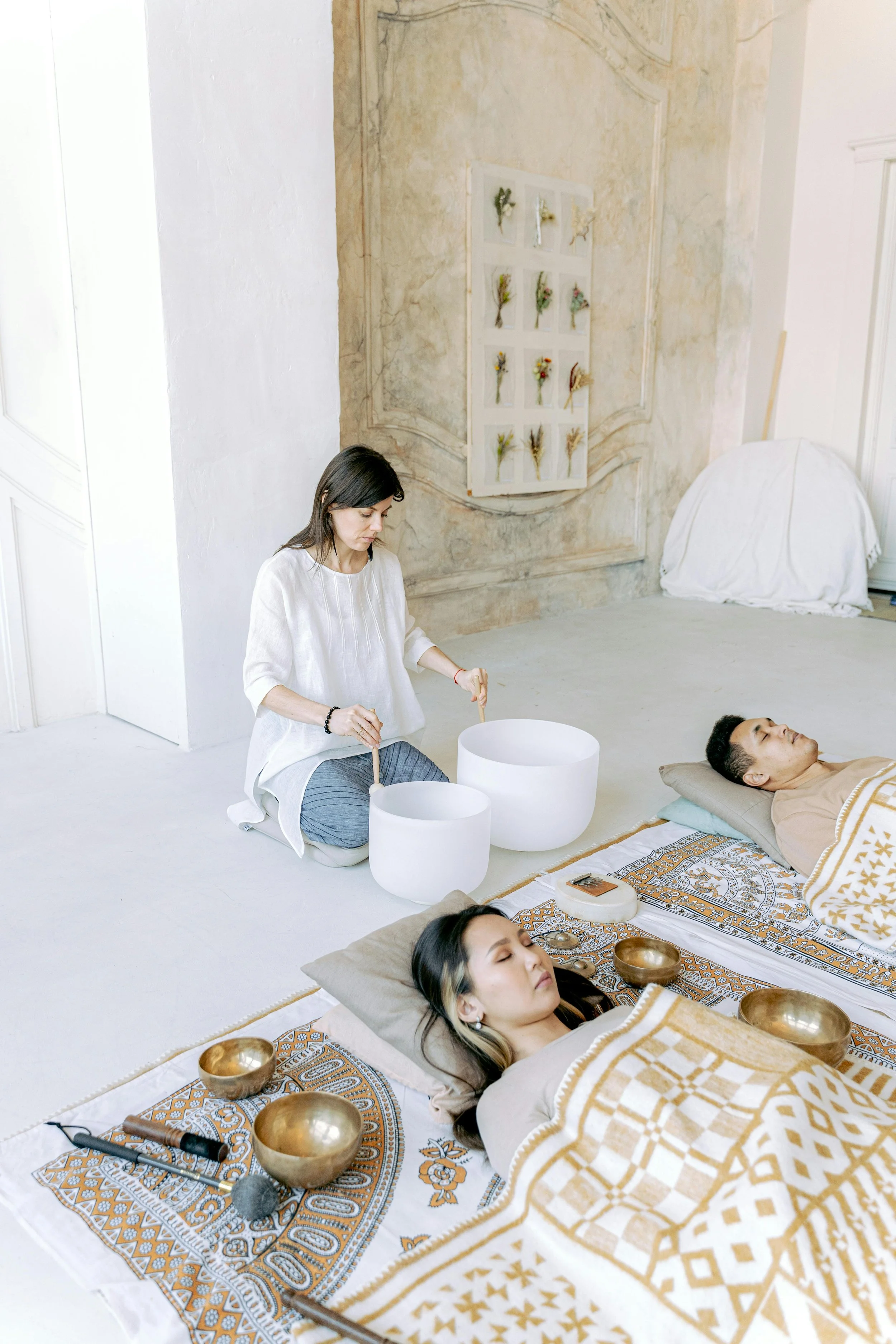 Three people lying on the floor with blankets over them, participating in a sound therapy session using singing bowls. A woman is seated nearby playing two large crystal singing bowls with mallets.