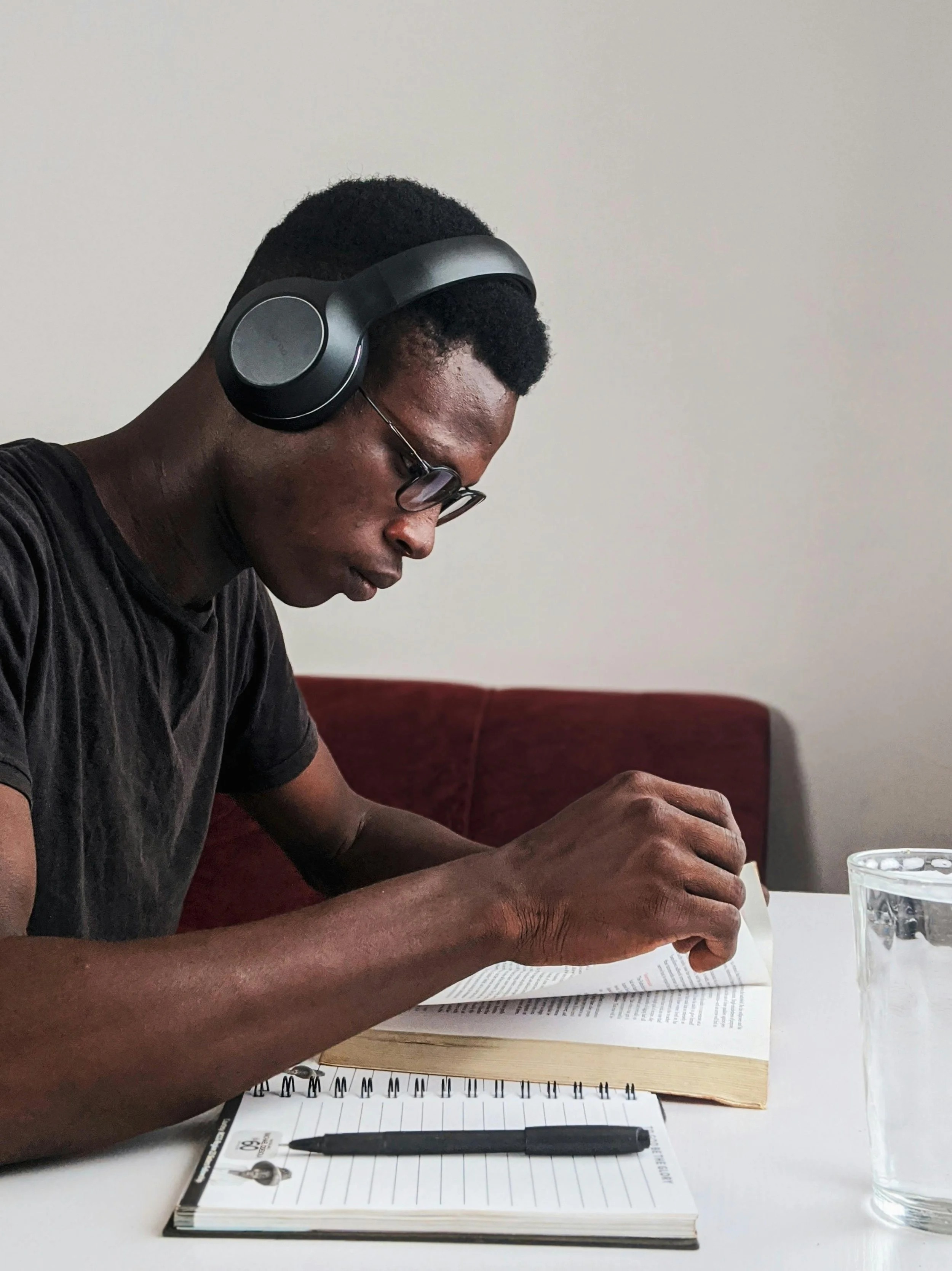 A young man wearing glasses and headphones is reading a book at a table. There is a glass of water and a notebook with a black pen in front of him.