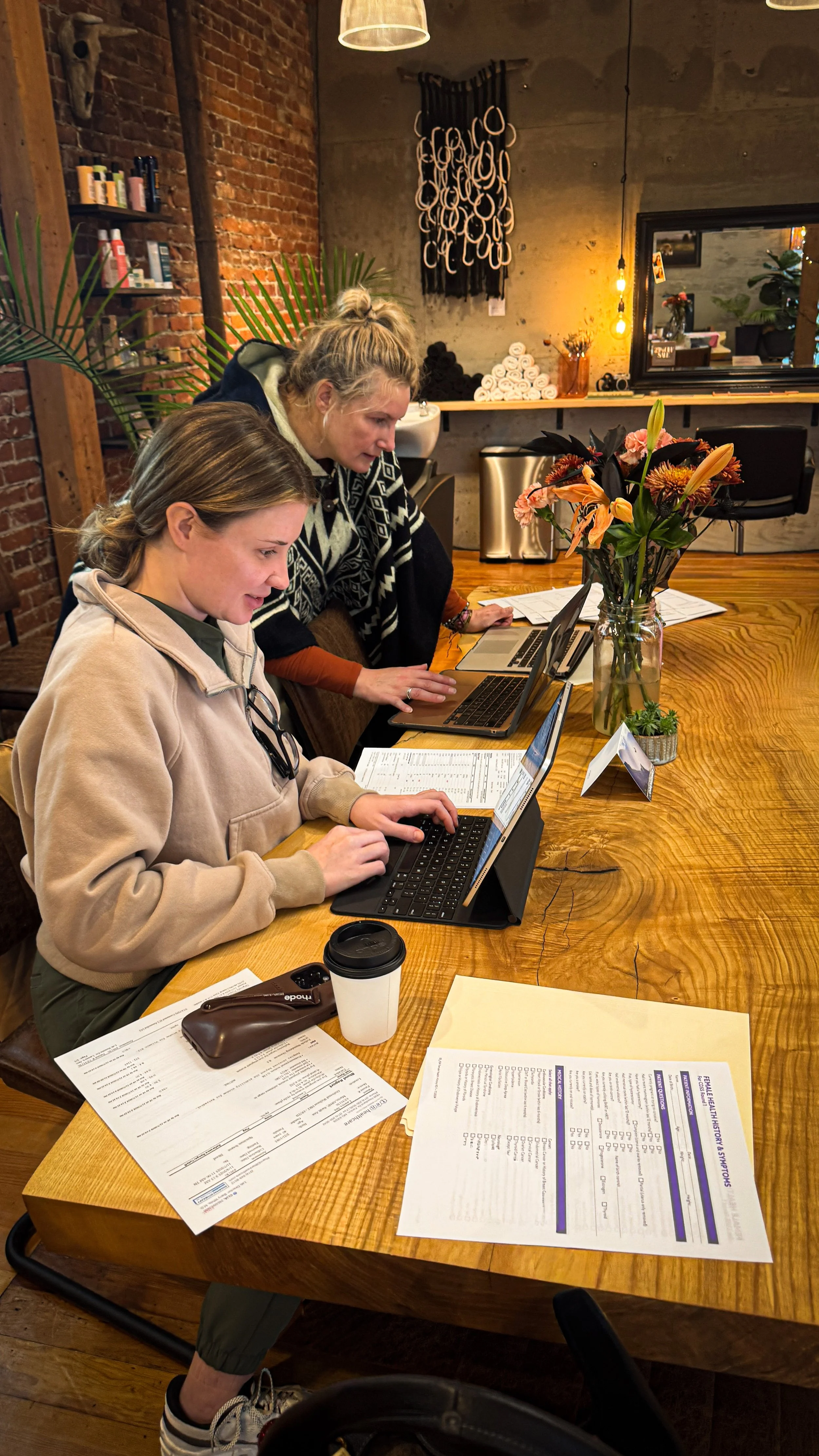 Two women working on laptops at a wooden table within Outbound Wellness . There are papers on the table and they are discussing Hormone Replacement Therapy for patients.