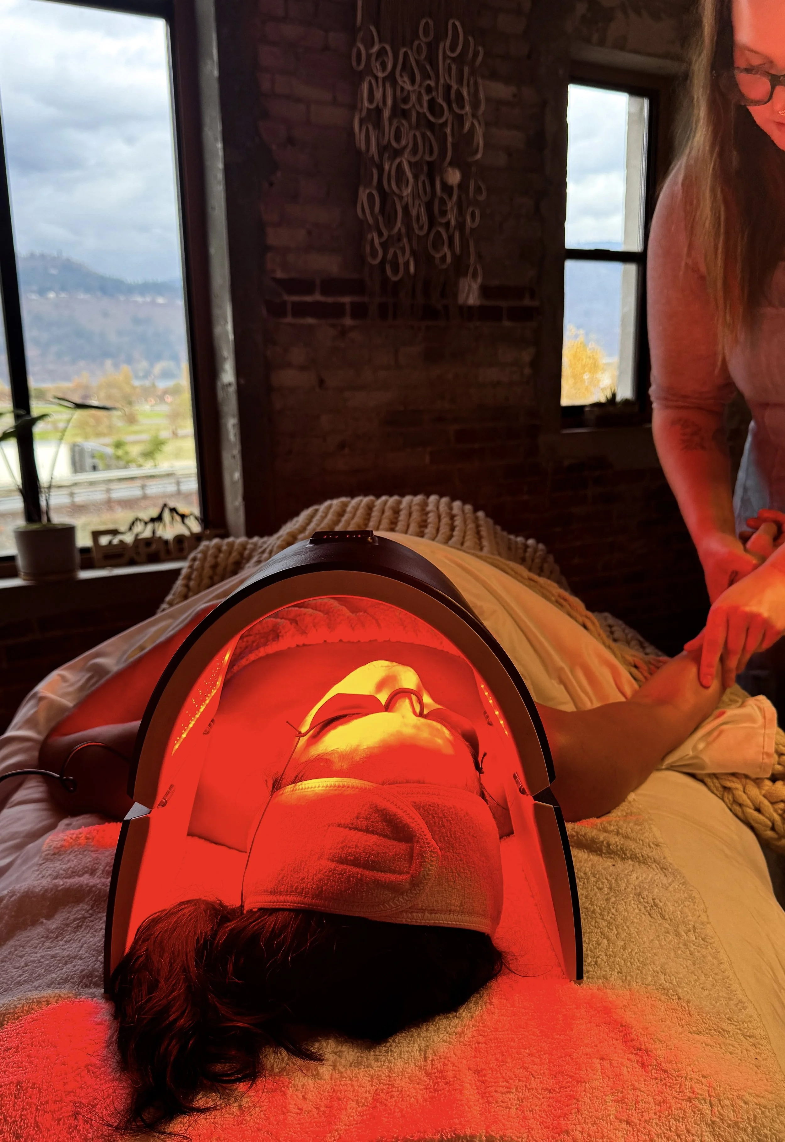 Person lying on a bed beneath a red LED therapy device, with a therapist massaging their arm, in a room with large windows and brick walls.