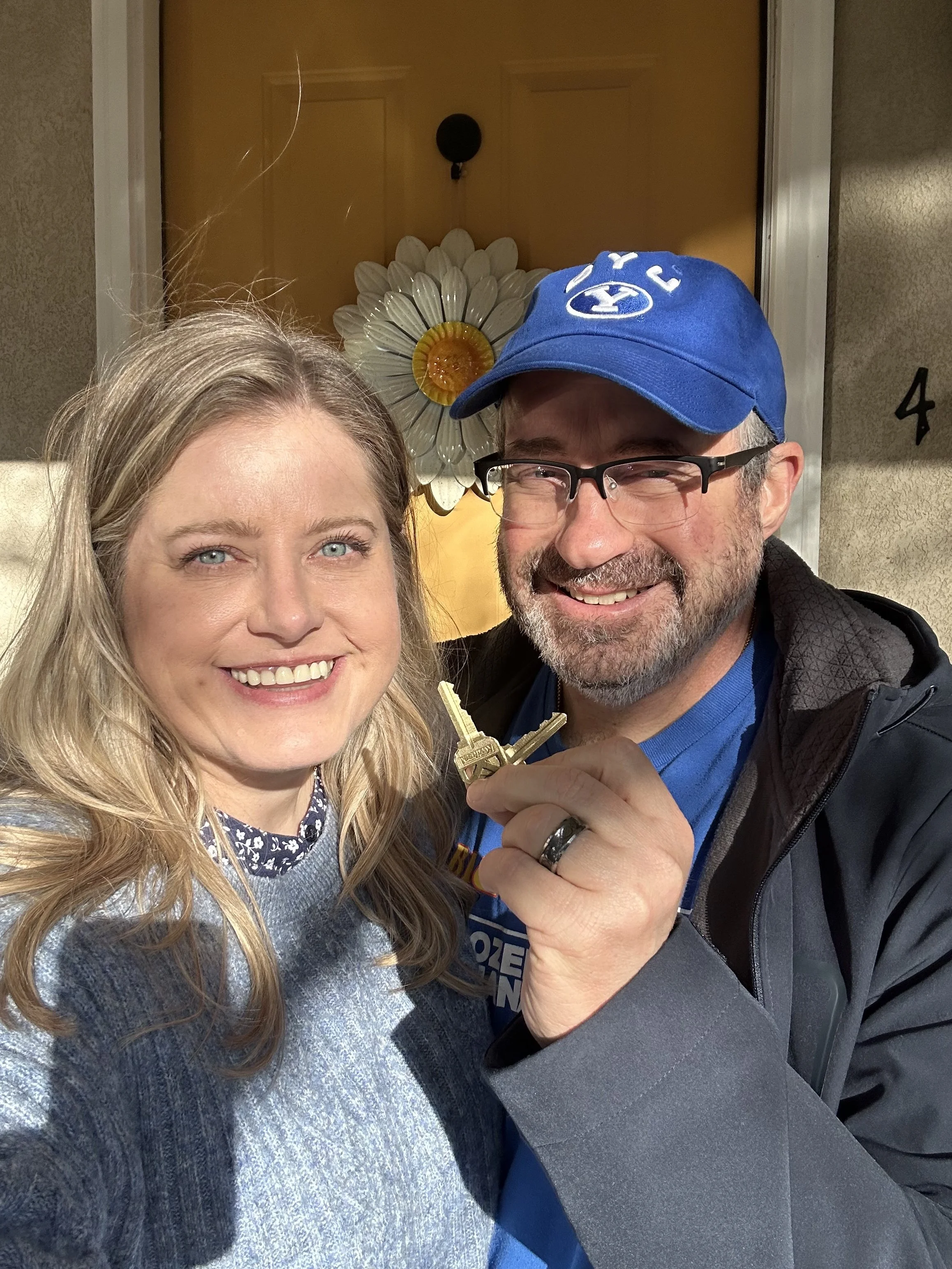 Close-up of smiling woman with blonde hair and a man wearing glasses, a blue cap, and holding keys, in front of a wall decor featuring a white flower-shaped ornament with a yellow center.