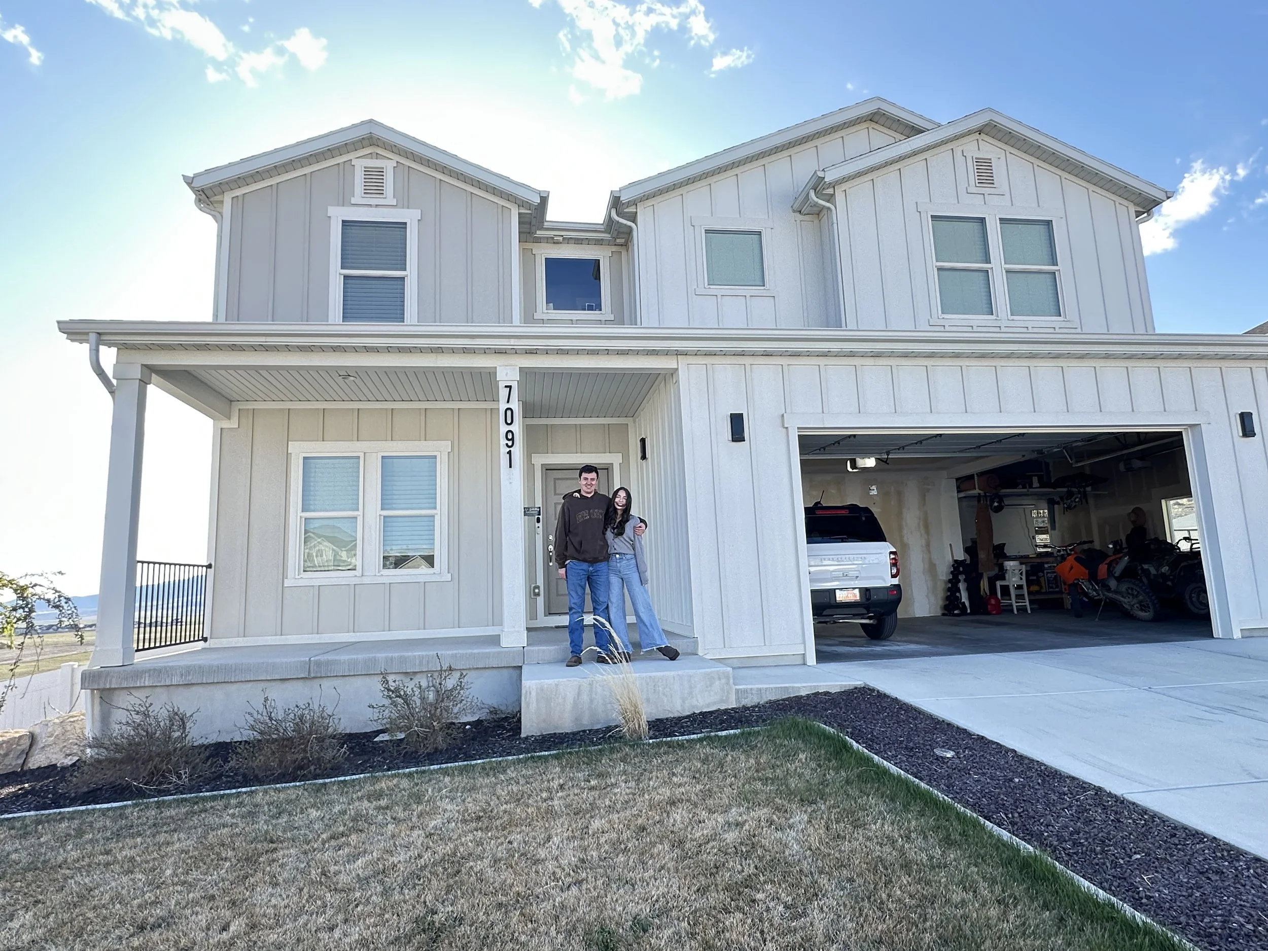 Two people stand in front of a modern two-story house with a gray exterior, white trim, and a garage. The house number is 70091. The yard has grass and a small garden bed with some plants, and a clear blue sky is overhead.
