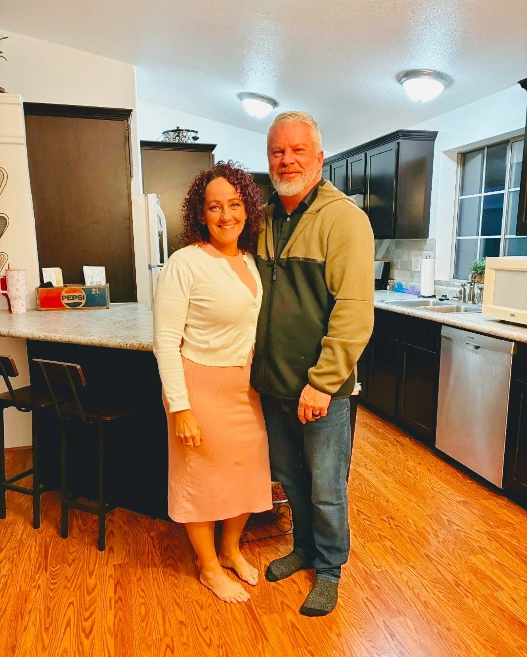 A happy couple standing in a kitchen, smiling at the camera. The woman has curly hair and is wearing a white cardigan and pink skirt, while the man has gray hair and beard, wearing a two-tone jacket and jeans.