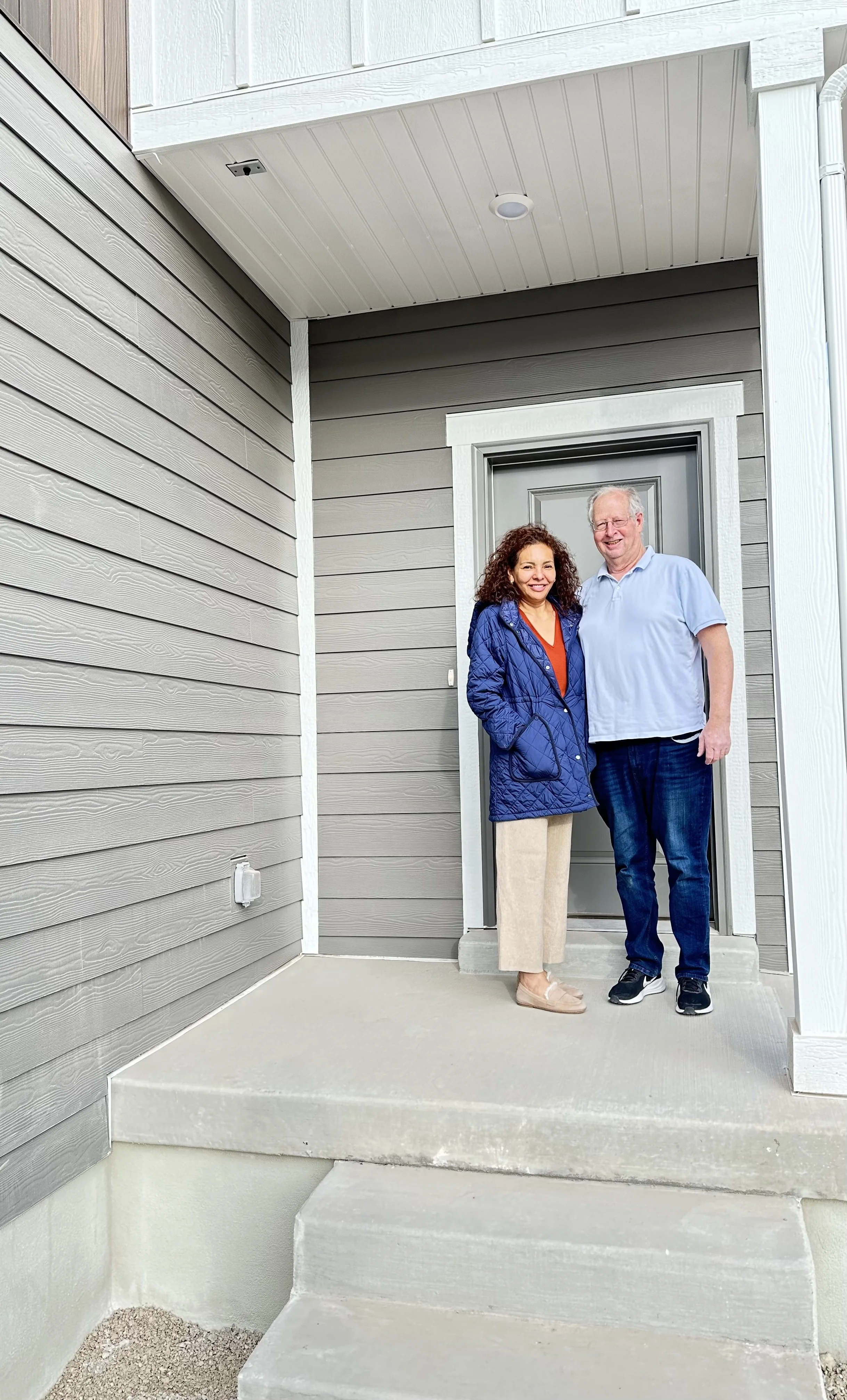Two people standing on a front porch, smiling, in front of a gray door, with the house's exterior featuring gray horizontal siding.