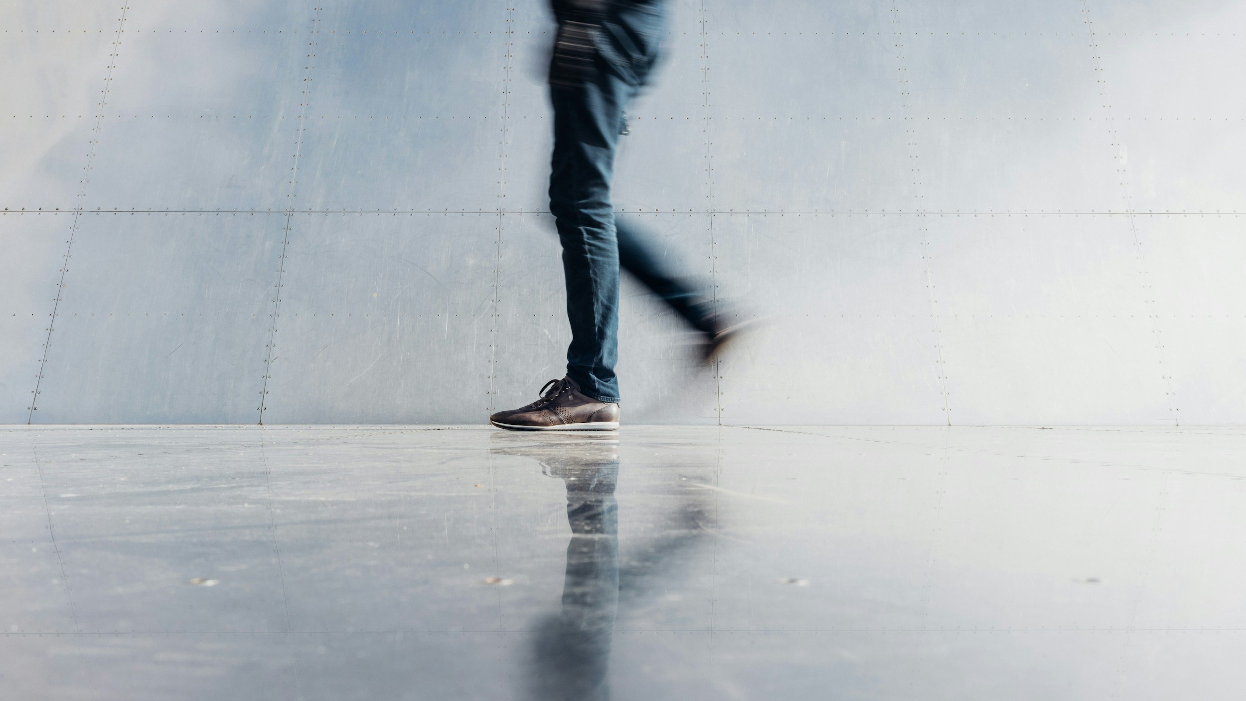 Person walking in a modern, metallic interior space, wearing dark jeans and brown shoes, with their reflection visible on a shiny floor.