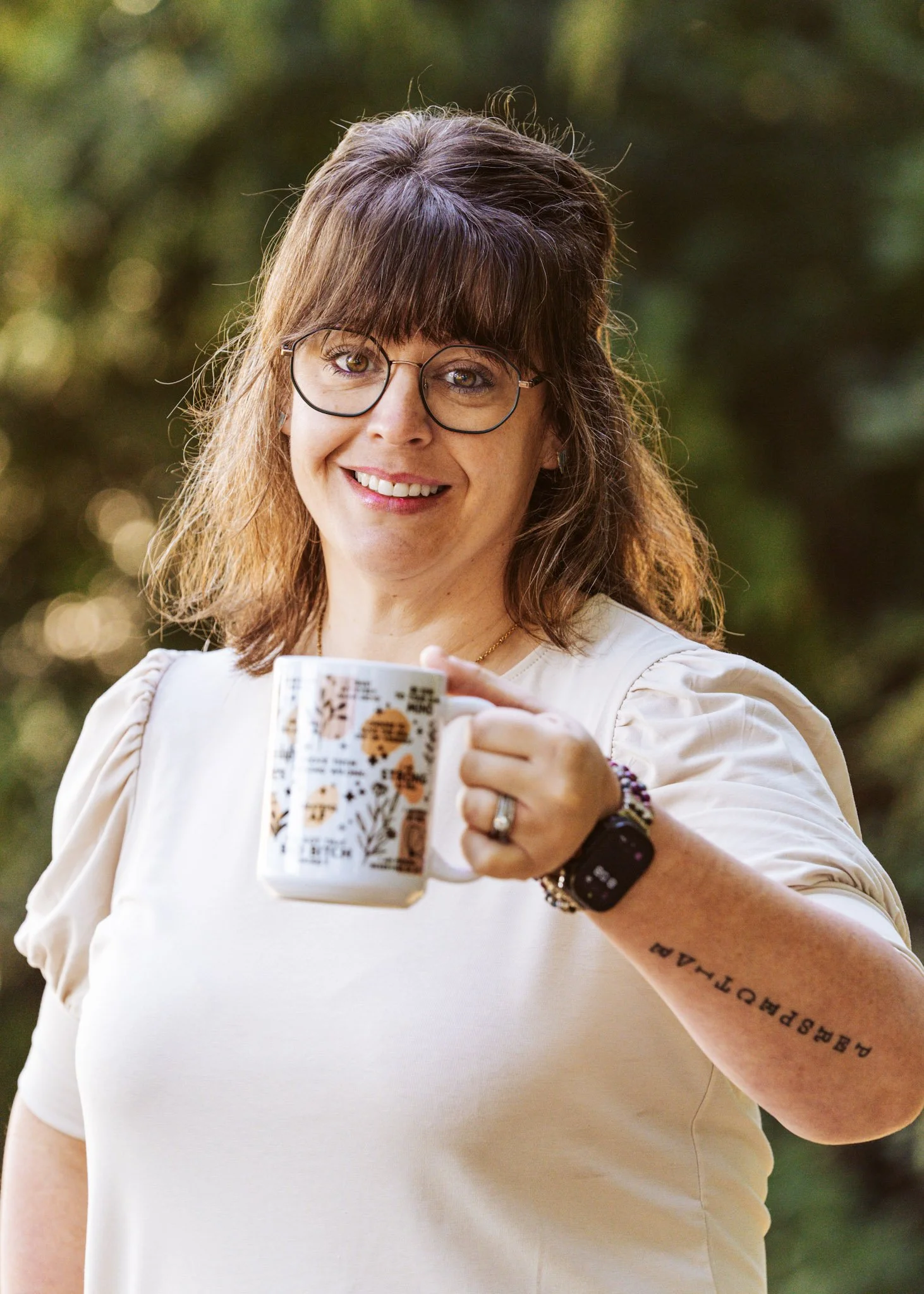 A woman with glasses smiling and holding a coffee mug outdoors, wearing a white shirt and a smartwatch.