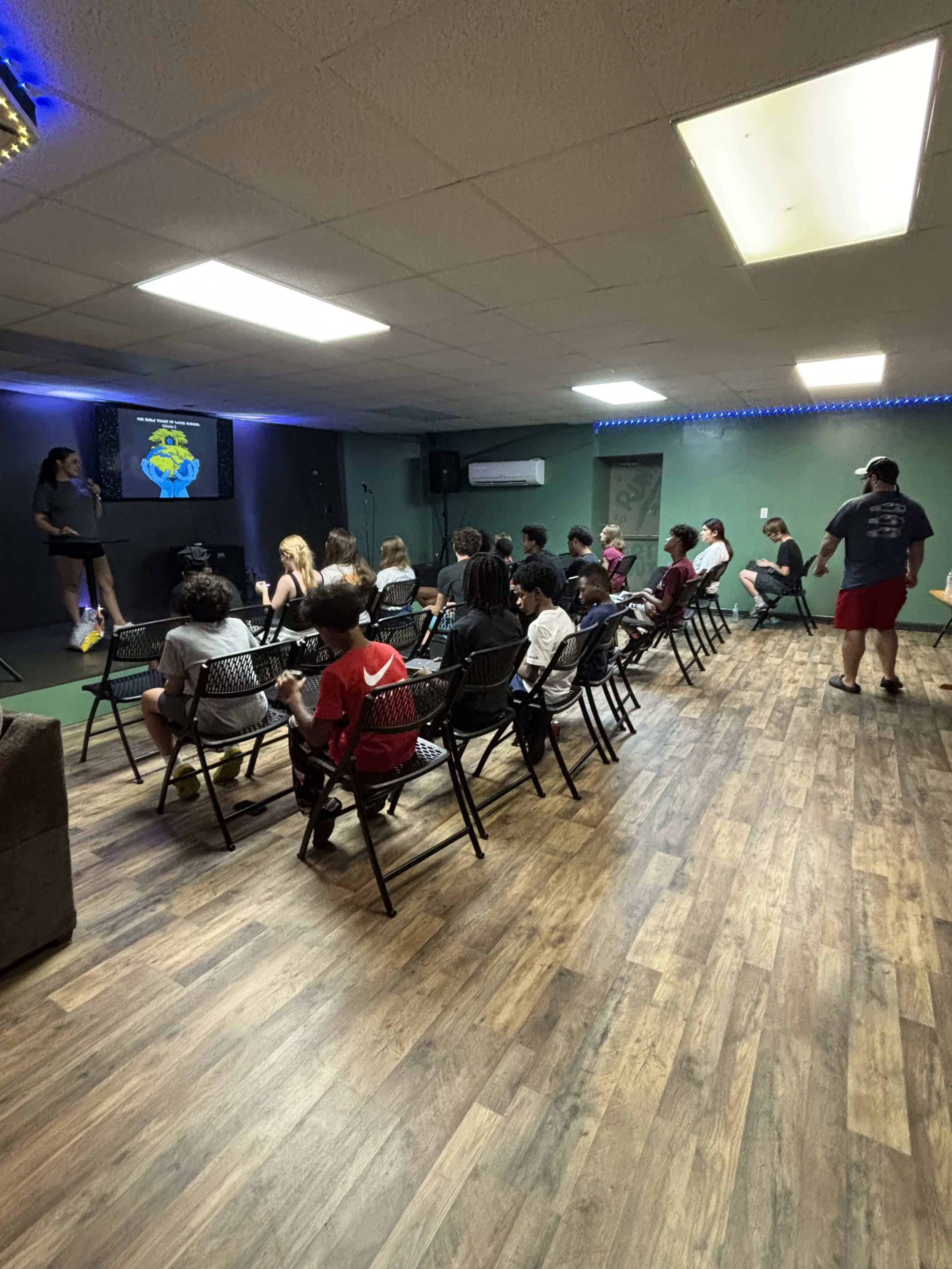 A group of people gathered around a table, looking at their phones, indoors with a sign that says 'Ginos Bar' in the background.