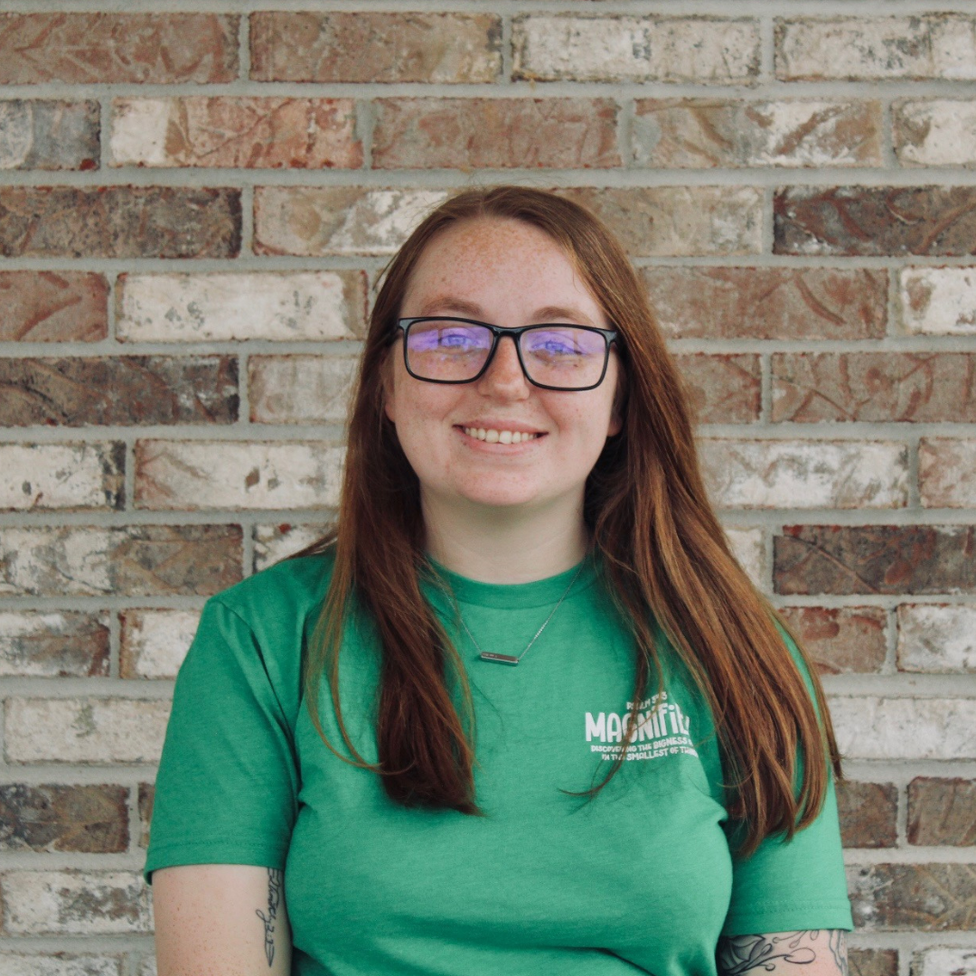 A woman with brown hair and blue eyes smiling, standing in front of a brick wall, wearing a black blouse with a blue and green floral pattern.