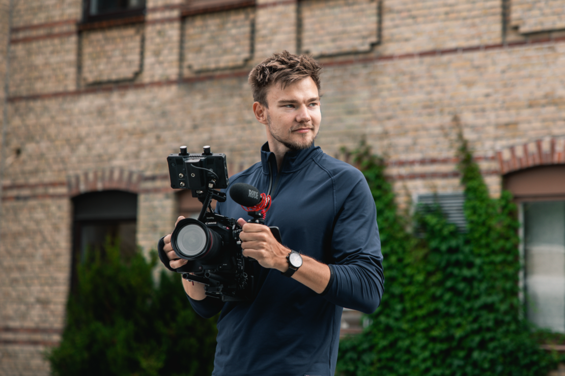 En ung man står utomhus framför en tegelvägg, håller i en professionell videokamera och fokuserar.