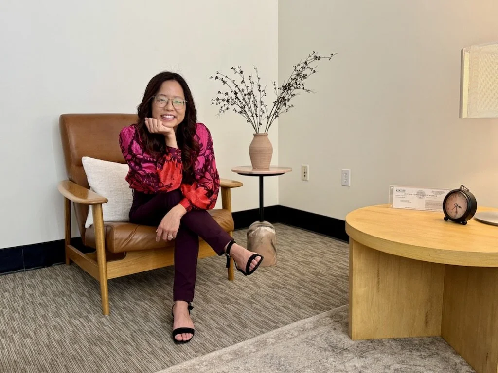 Woman sitting on a wooden chair in an office, smiling, wearing glasses, a pink and red floral blouse, and black sandals, with a white pillow behind her, a table with a vase of dried branches, and a round wooden desk with a clock and a paper on it.