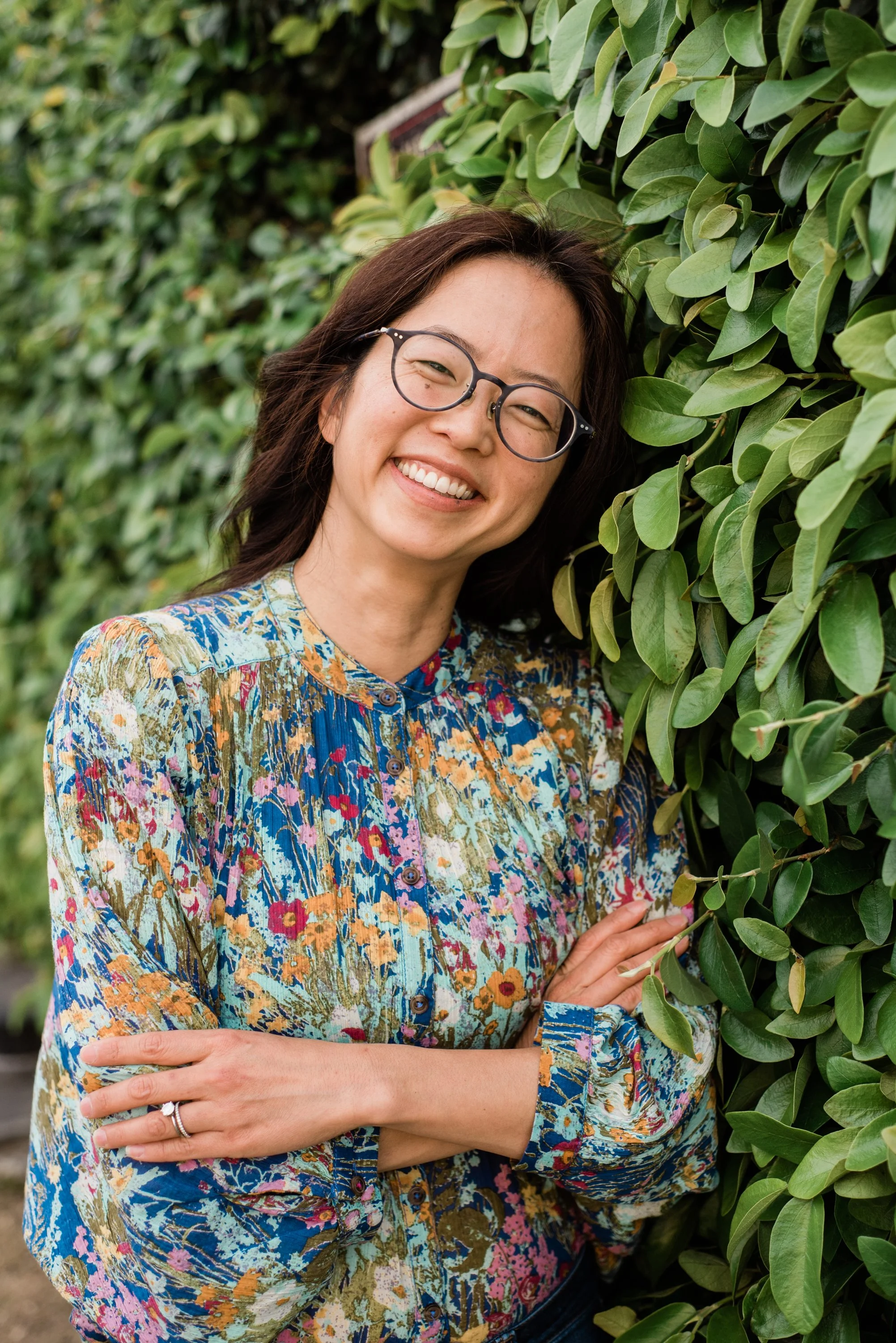 A woman with glasses and long dark hair smiling, standing next to green leafy bushes, wearing a colorful floral blouse.