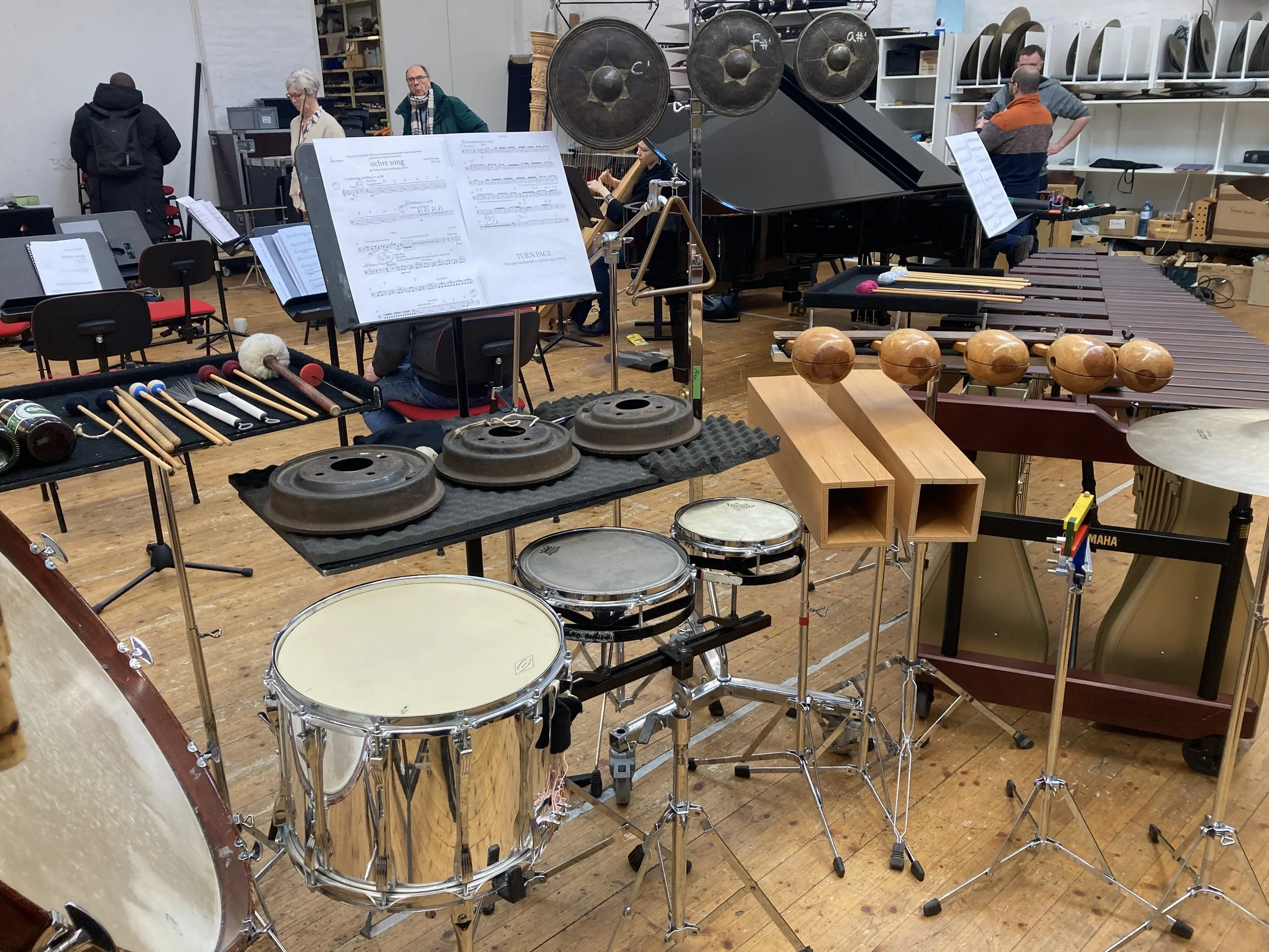 A percussion setup including 3 brake drums, 3 gongs, 2 log drums, 5 temple blocks, 3 toms, a triangle, a ratchet, a marimba, and the printed music. Several musicians are also visible, moving around the room, preparing for the rehearsal.