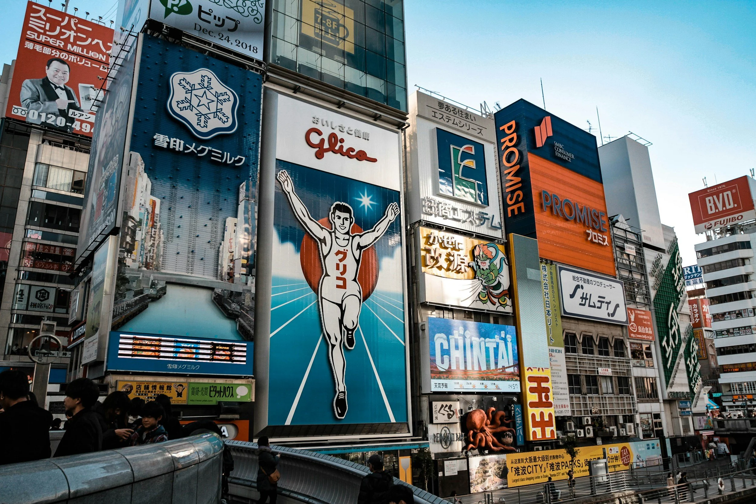 City scene with numerous colorful billboards and advertisements on tall buildings, including a large running figure and various text in Japanese and English. Pedestrians are visible at the bottom of the image.