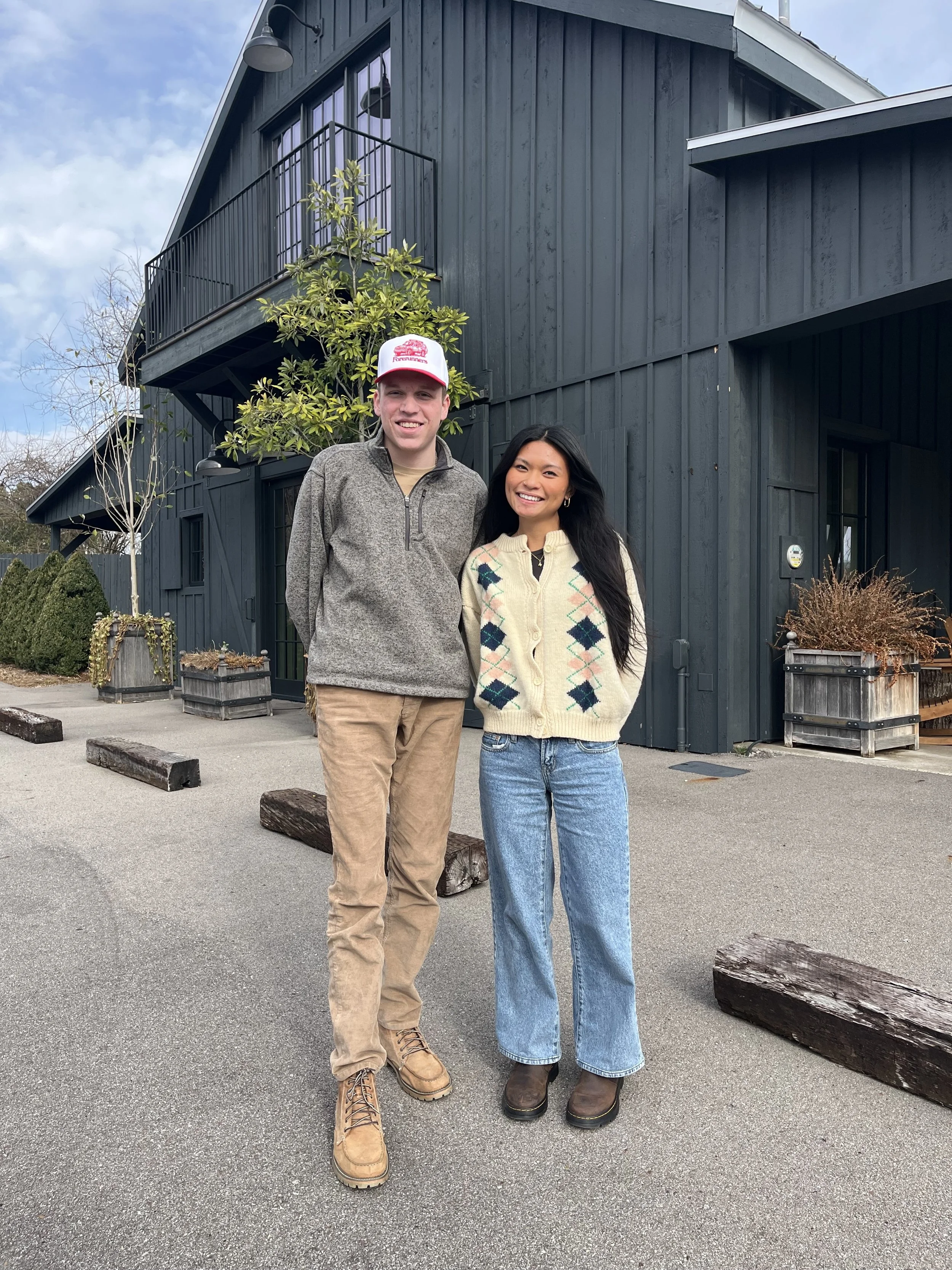 A young man and woman standing outdoors in front of a dark blue building, smiling at the camera. The man is wearing a gray sweater and tan pants with tan boots, while the woman is wearing a cream-colored sweater with diamond patterns and wide-leg jeans with brown boots.
