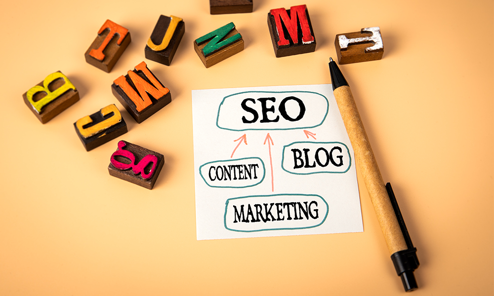 A piece of paper has  flow chart with SEO at the top and content, blog and marketing point towards it. Next to it is a pen and  stamps with letters of the alphabet are scattered above the flowchart.