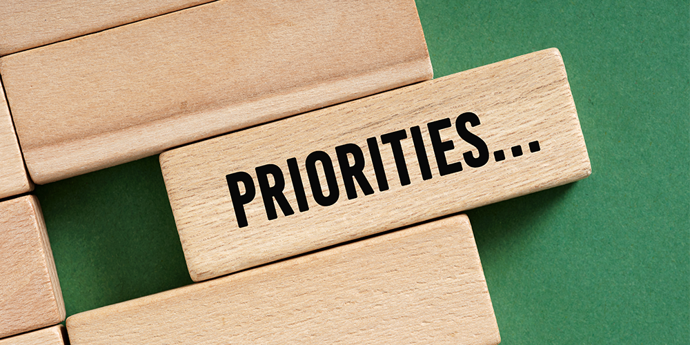 A cluster of wooden tiles rest on a green background. One tile is sliding out from the rest with the word "Priorities..." stamped on it.