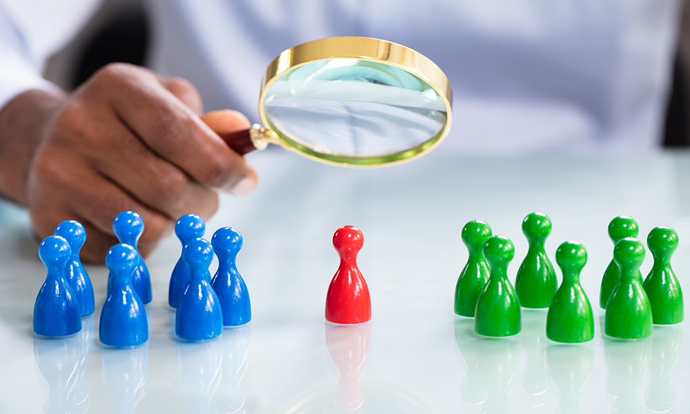 3 groups of small board game pieces are on a white table. There is a blue group of 7 on the left. A group of 7 green pieces on the right and one red piece in the middle. Above the game pieces, a man holds a magnifying glass over the red piece.