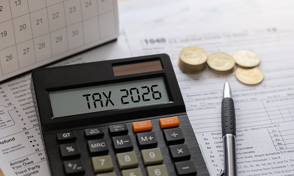 A calculator sits on a pile of tax documents with a pen and a stack of quarters. The calculator says, "Tax 2026" on the screen.