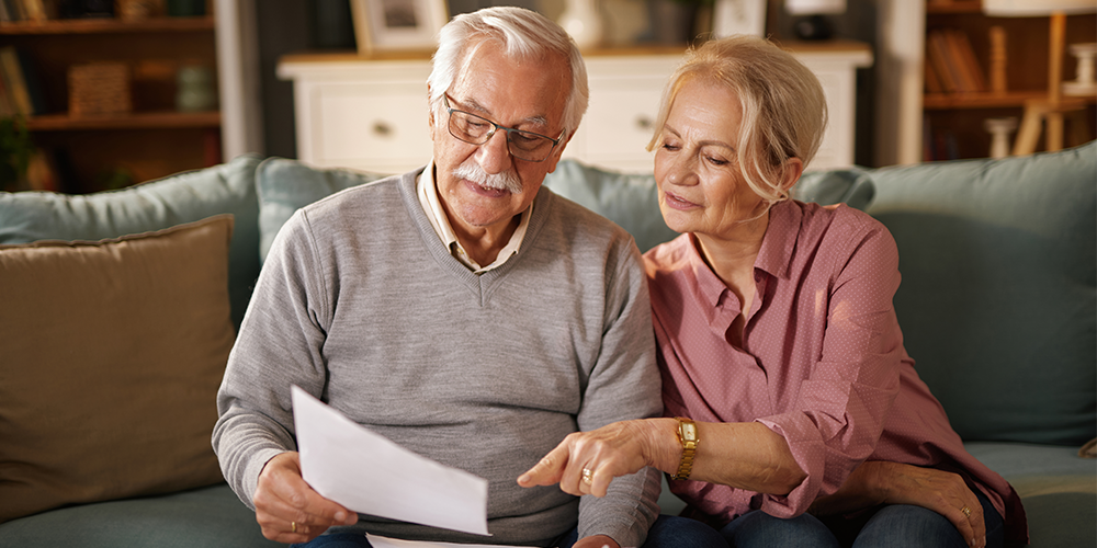An older caucasion couple sit on a sofa and are looking at a piece of paper.