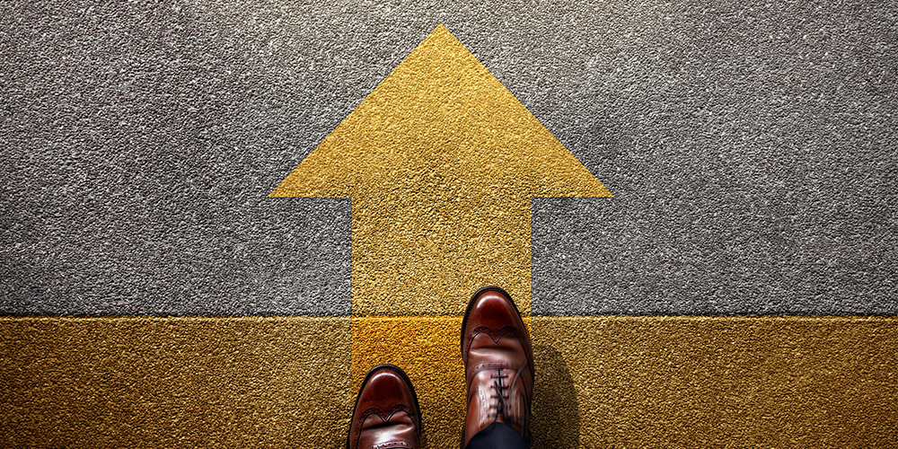 A pair of feet in men's dress shoes stand at the bottom of a yellow arrow that's painted on the street. The arrow is pointing up.