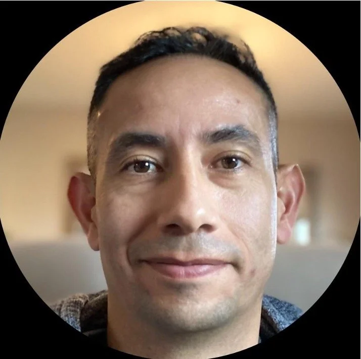 Close-up of a smiling man with short dark hair, brown eyes, and a light complexion in an indoor setting.