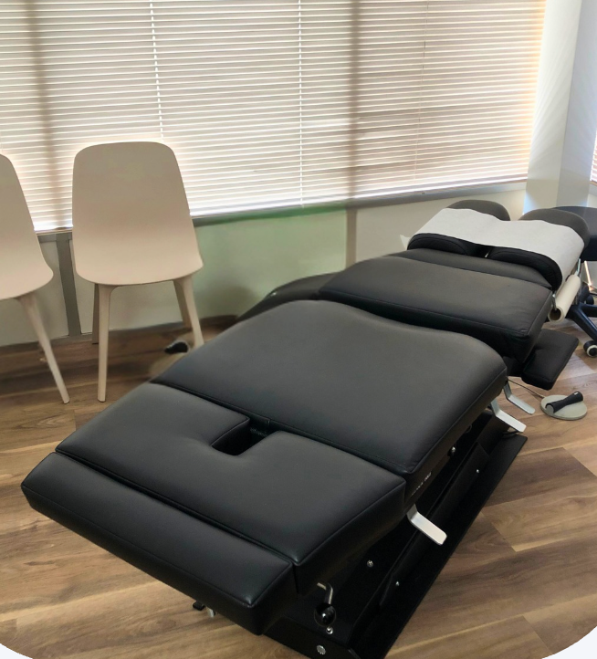 A comfortable chiropractic table in a chiropractic treatment room with chairs and closed blinds in the background.