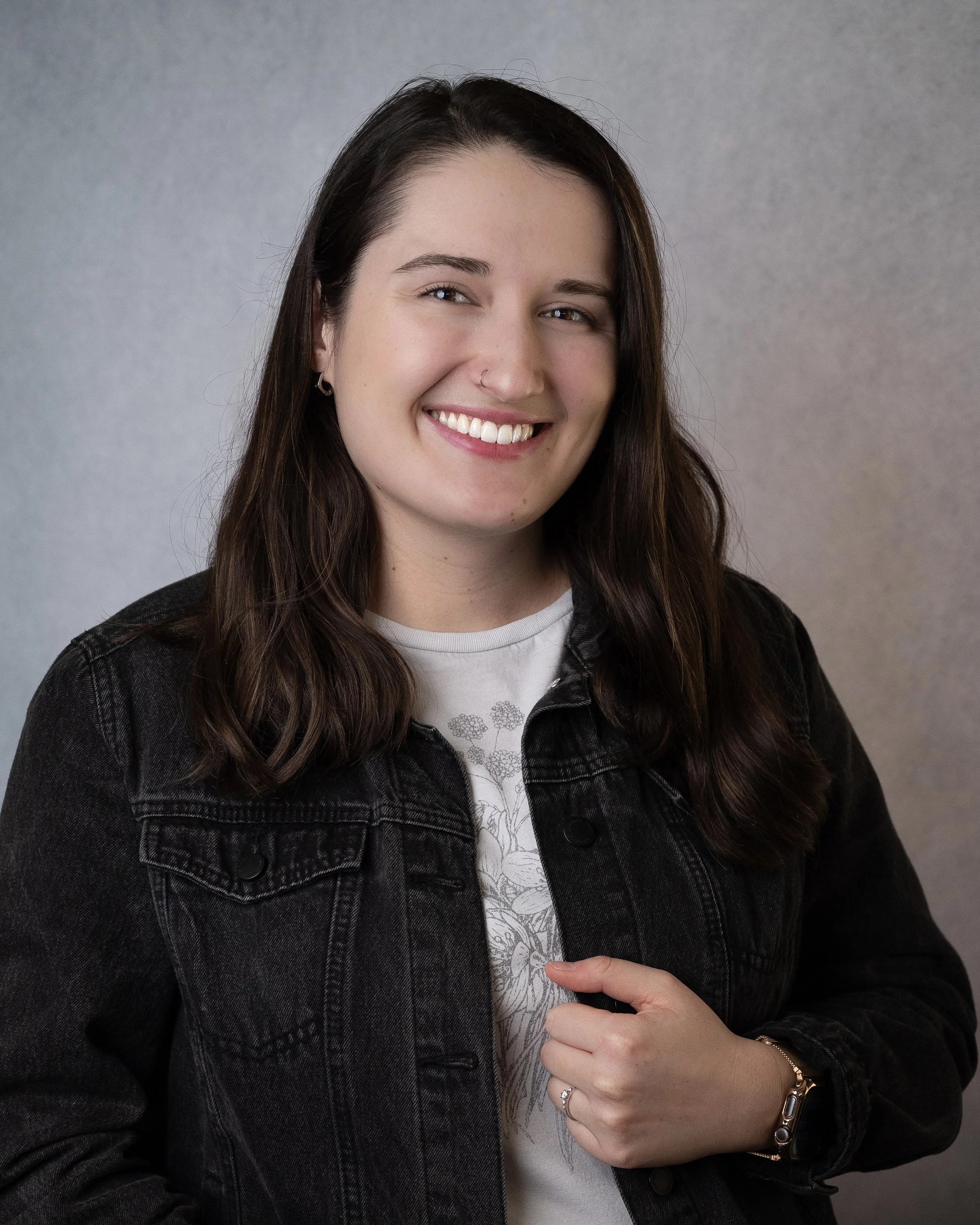 A young woman with dark brown hair, light skin, and a nose ring, smiling in front of a plain green background, wearing a reddish-orange embroidered top.