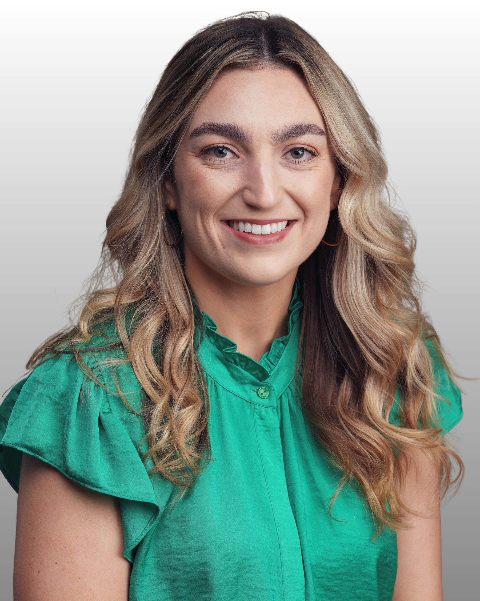 A woman with long, wavy blonde hair and light skin, smiling, wearing a green blouse, against a plain, muted background.