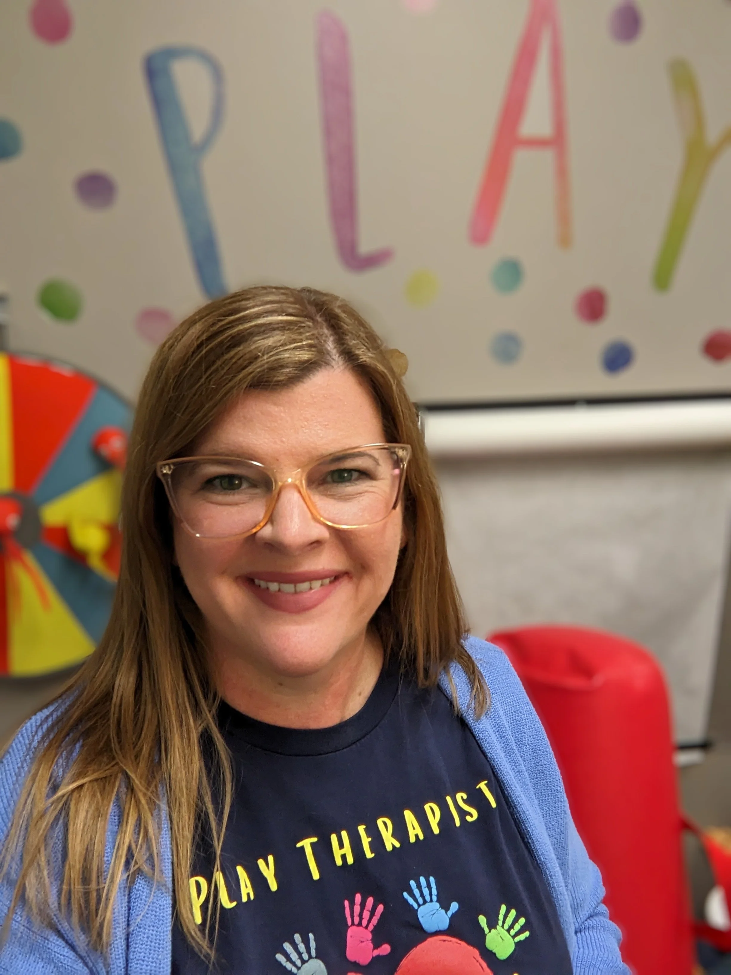 A therapist with long hair in a play room with a shirt that says "Play Therapist"