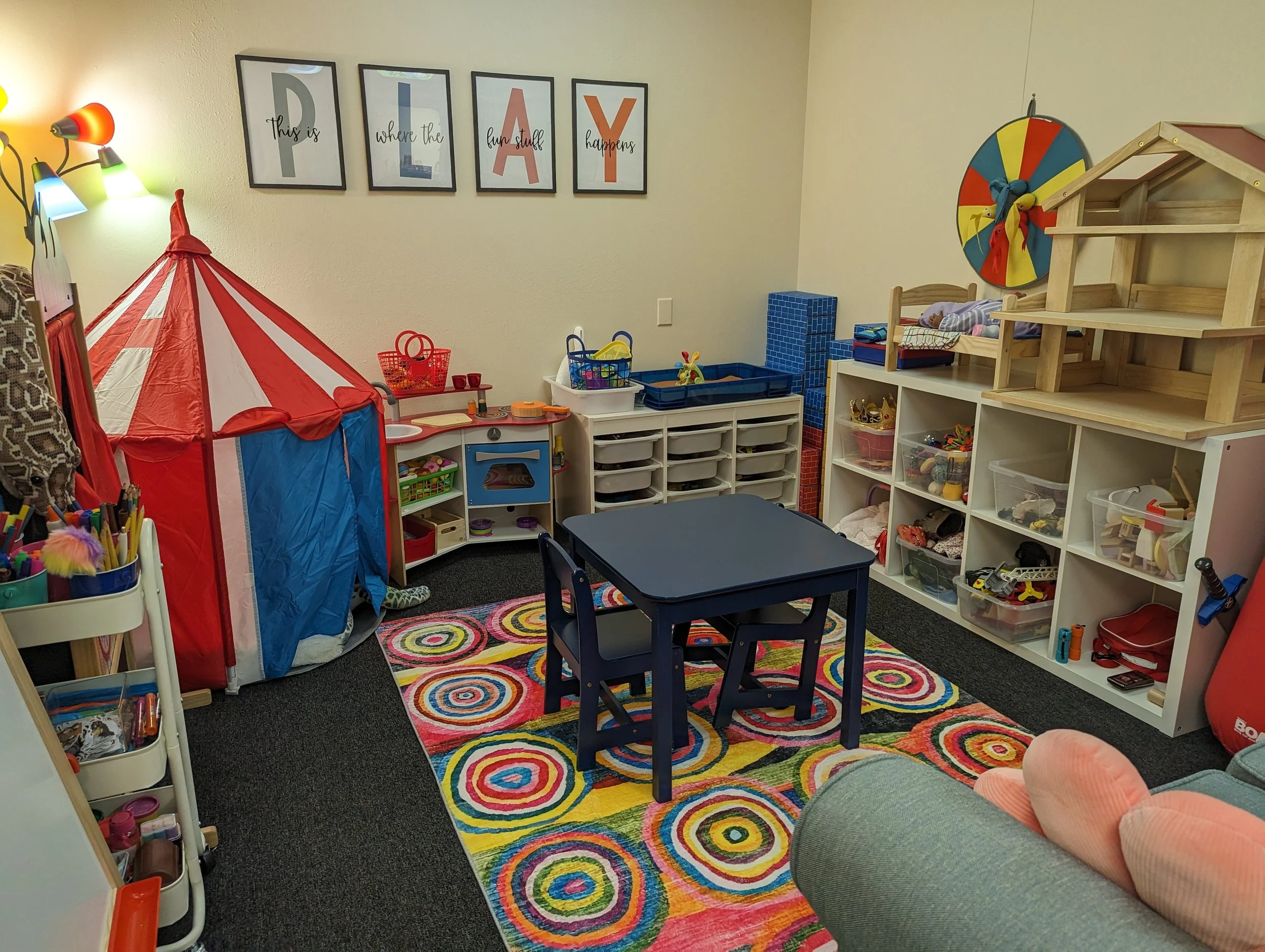 Children's playroom with a colorful circular rug, small table and chairs, a red and white play tent, shelves filled with toys, and wall decor spelling out 'PLAY' and 'This is where the fun stuff happens'.