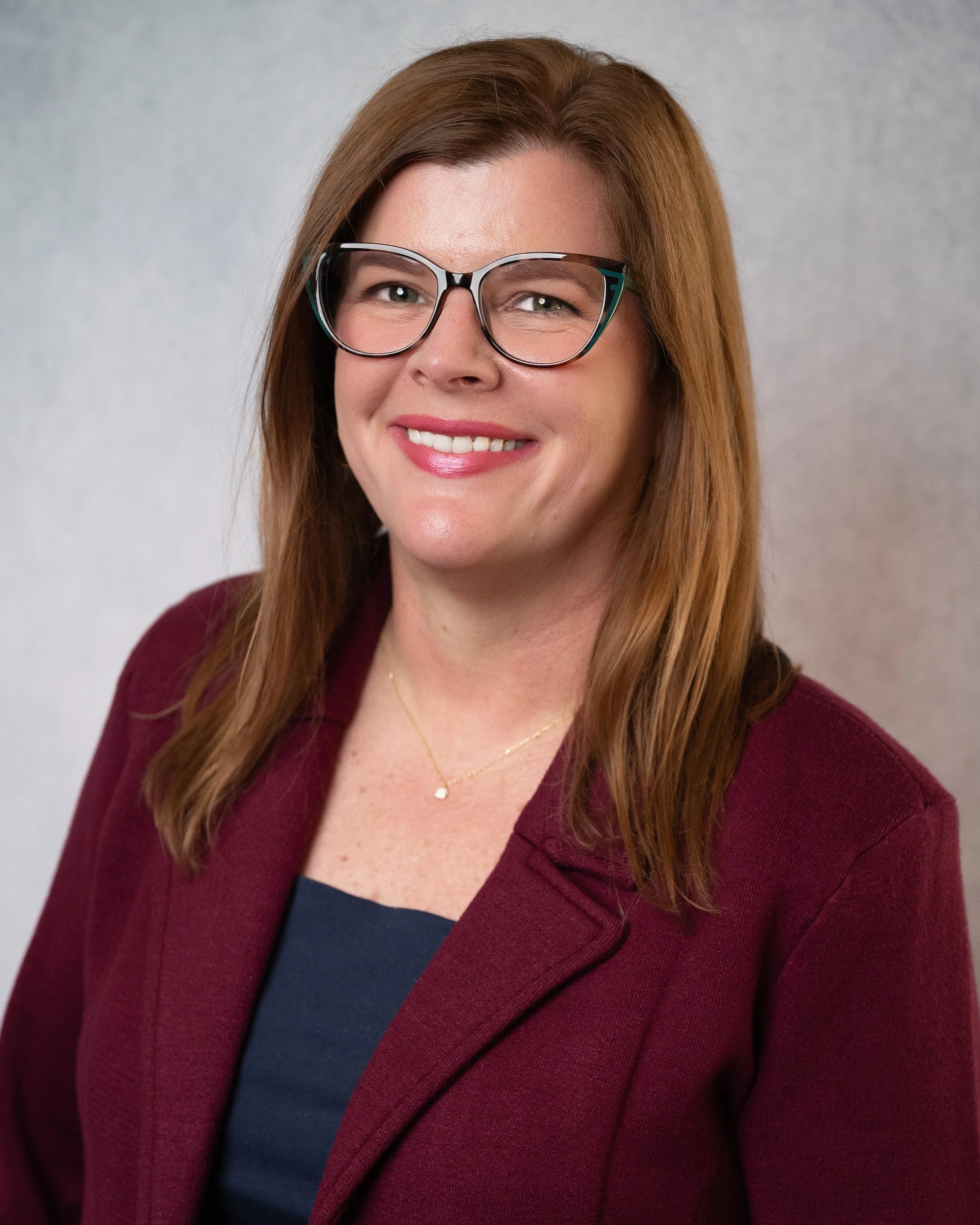A woman with light skin, long brown hair, wearing clear-framed glasses and a red shirt, smiling against a neutral background.
