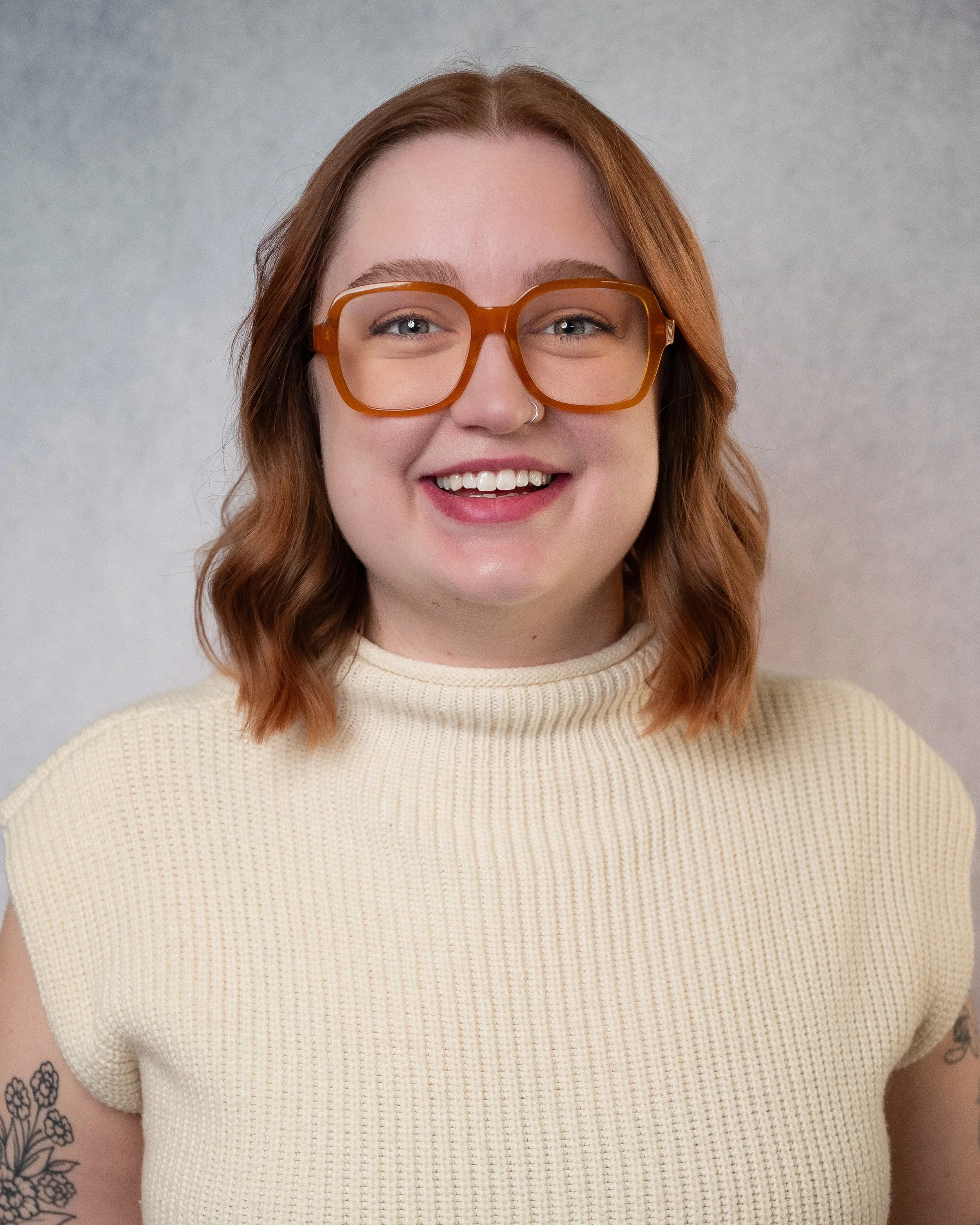 A young woman with shoulder-length wavy brown hair, wearing glasses and a nose ring, dressed in a light green ruffled top, smiling at the camera against a neutral background.
