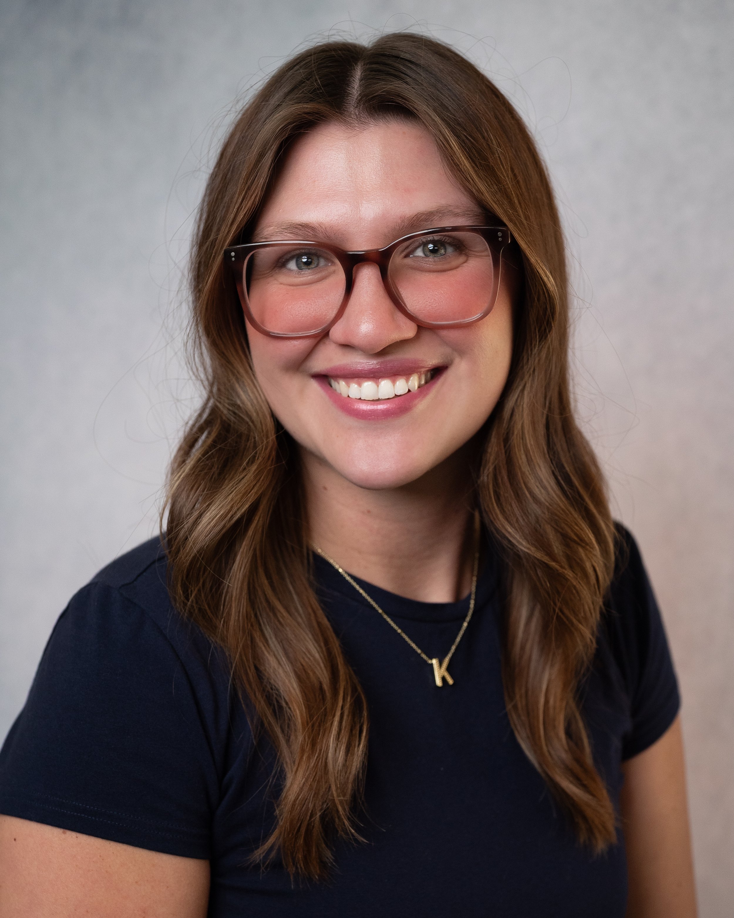 A woman with shoulder-length brown hair, glasses, and a white t-shirt smiling in front of a beige wall. The t-shirt has the text 'Kindness Knows No Limits Love Everyone' printed on it.