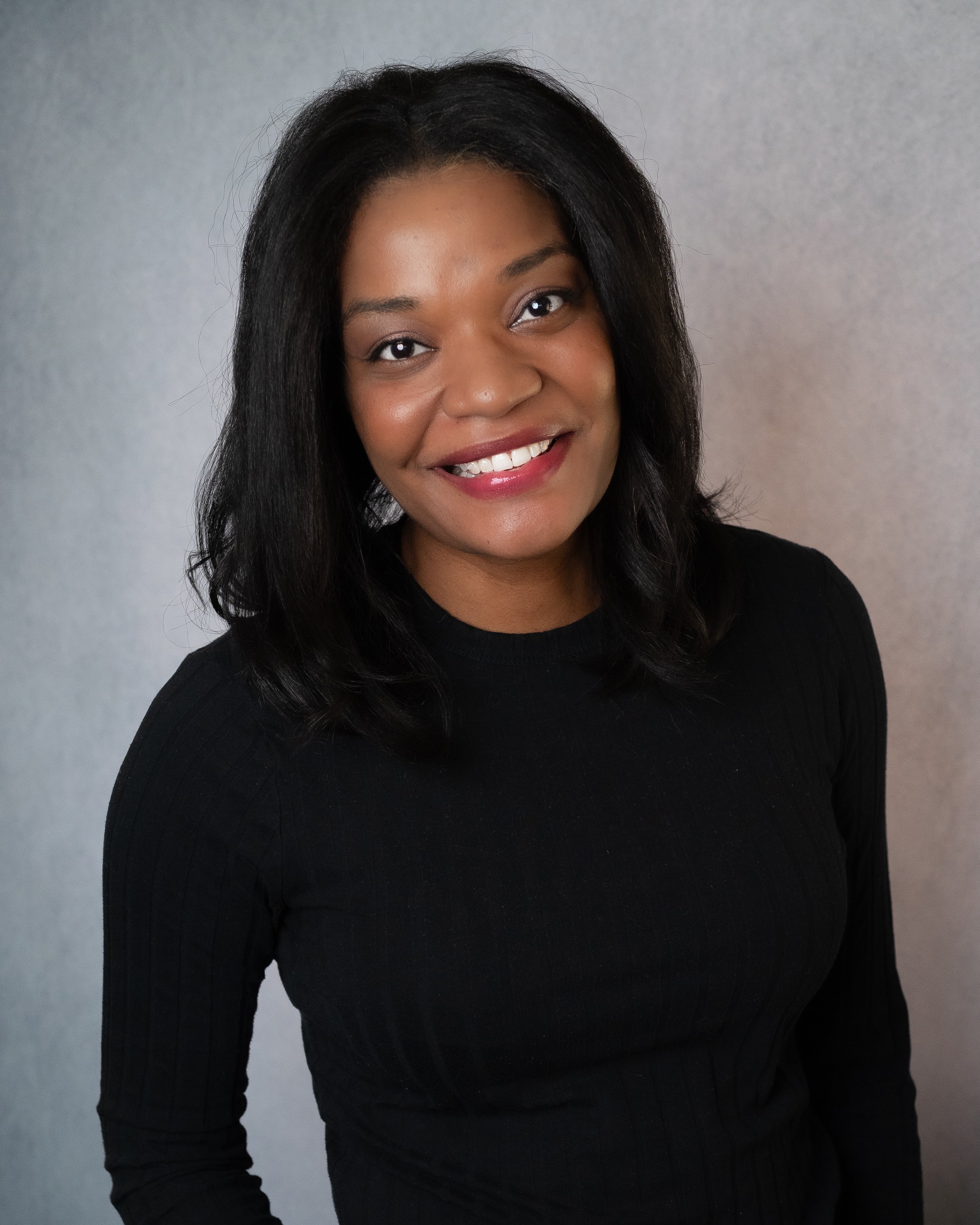 A woman with black hair smiling, wearing a white shirt and green sweater, against a brownish-gray background.